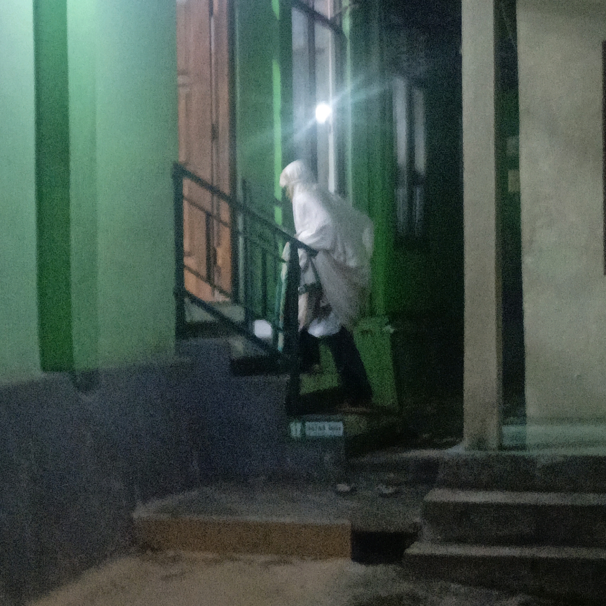 The image captures an outdoor scene at night, likely in Salatiga, Indonesia, given the presence of Arabic script on a sign. A person dressed in white, possibly a religious garment, is ascending a set of stairs. The stairs are bordered by a black metal railing and lead up to a doorway. The building has a green facade and large windows. A bright light is visible, potentially a streetlamp or interior lighting, illuminating the area. The ground surface appears to be paved. The overall lighting suggests it is nighttime or a dimly lit area. The sign with Arabic script, reading "17", is partially visible on the wall near the stairs, indicating a possible address or location marker. The scene is rendered with a grainy quality, characteristic of low-light photography.