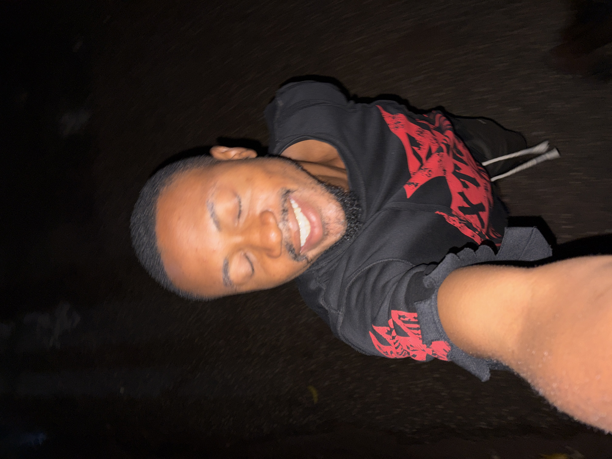 The image captures a selfie of a man, set in Jos, Nigeria, taken at what appears to be night due to the extremely dark background. The man is positioned from a low angle, possibly lying down or leaning back, with his arm outstretched towards the camera. He is smiling widely, showing his teeth, with his eyes closed, conveying a sense of joy or amusement. He has a beard and short, dark hair.
He is wearing a black hoodie featuring a prominent red design on the chest and sleeve. This design appears to be stylized text or an emblem, though it is not clearly legible. White drawstrings are visible near his left shoulder. The background is a very dark, indistinct surface, possibly asphalt or rough ground, with minimal detail, emphasizing the subject.