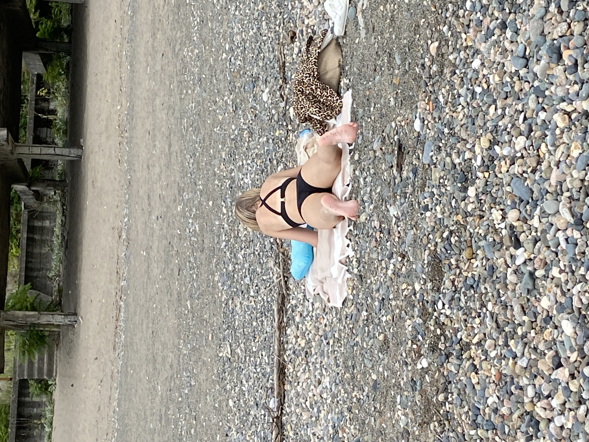 A woman with blonde hair is lying on a beach towel on a pebble-covered shore. She is wearing a black bikini and is positioned facing away from the camera. A blue pillow is placed behind her head. To her right, a leopard-print garment is draped over a bag, and a pair of white sneakers are visible next to it. The background shows a sandy area with concrete structures and vegetation, consistent with the urban coastline of Sokhumi, Abkhazia.