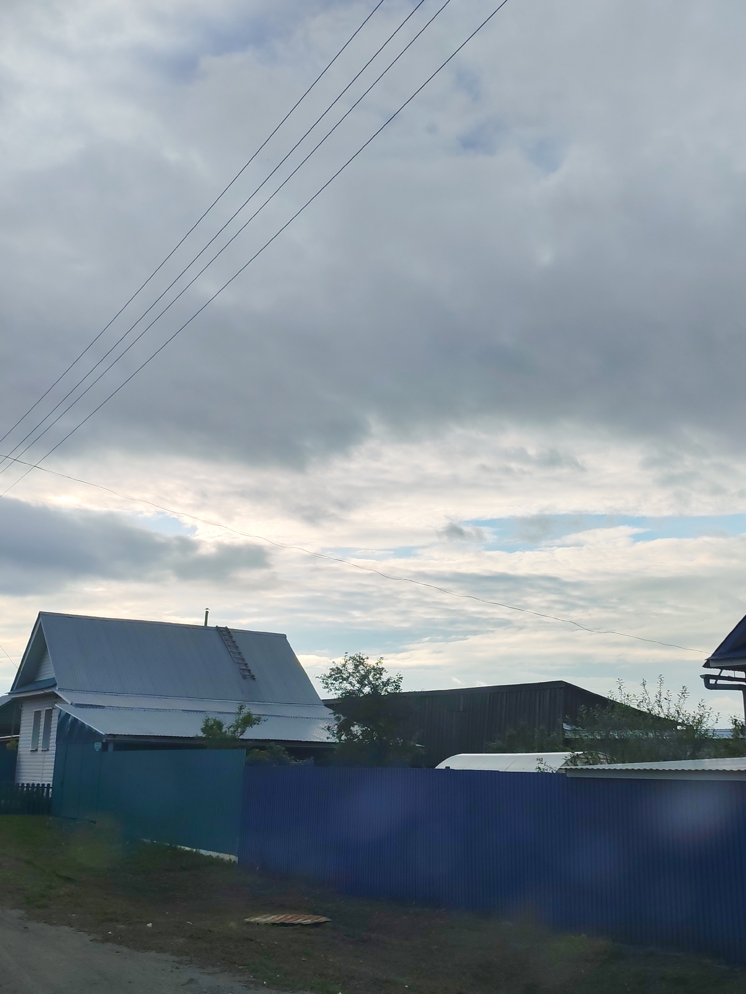 The image depicts a rural or semi-rural scene in Malaya Bod'ya, Russia, under an overcast sky with patches of visible blue. The upper two-thirds of the frame are dominated by gray and white clouds, with a clearer section of blue sky and brighter light appearing towards the mid-right. Multiple thin black power lines traverse the sky diagonally from the upper left, while a thicker, single power line stretches horizontally across the middle of the sky, slightly above the visible structures.
In the mid-ground, a white two-story house with a steeply pitched gray metal roof stands on the left. A wooden ladder is propped against the right slope of its roof, extending from the eave towards the ridge, where a small chimney or vent pipe is visible. An additional, lower gray metal roof extends from the main house. A corrugated metal fence, predominantly dark blue but appearing dark teal on its left section, runs across the mid-ground, separating the foreground from the properties beyond.
To the right of the fence, a dark, presumably wooden, rectangular building with a dark roof is visible behind dense green foliage. A lighter-colored, rounded structure, possibly a covered vehicle or greenhouse, is partially visible behind the fence and in front of the dark building. The edge of another roof with a gutter system is visible in the far upper right corner.
The foreground consists of a dirt or gravel path/road, with green grass and darker vegetation bordering it. A circular metal grate or manhole cover is partially visible on the ground in the lower-right quadrant. No individuals or animals are present in the scene.