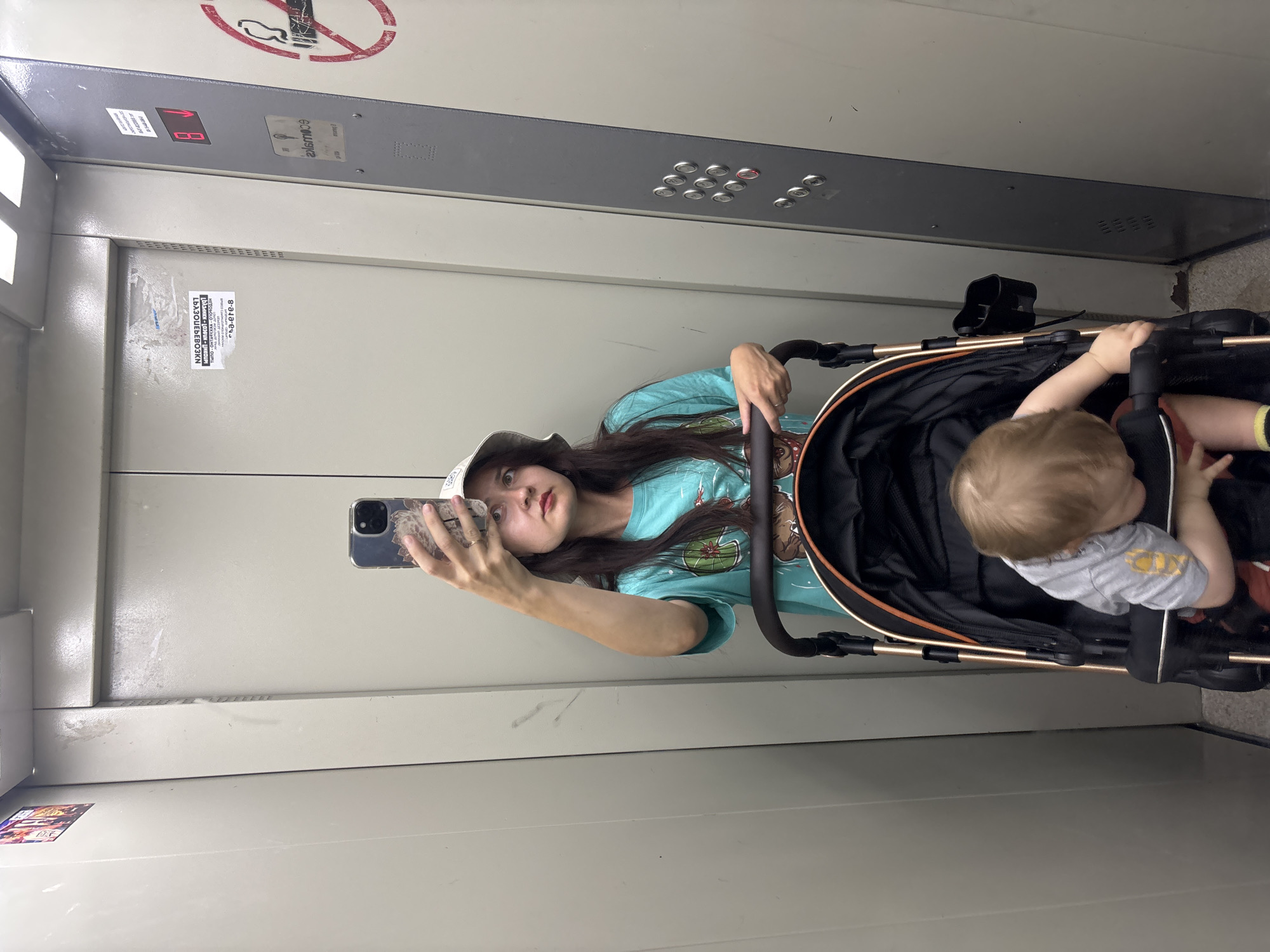 A woman is taking a mirror selfie in an elevator in Naberezhnye Chelny, Russia. She is wearing a light-colored hat and a turquoise t-shirt with a graphic print of water lilies. Her left hand holds a smartphone, and her right hand is on the handle of a stroller. A baby, wearing a grey t-shirt, is visible in the stroller. The elevator walls are grey and metallic. On the wall, there is a sticker with Cyrillic text that reads "ДЬАЗОЦЕЬЕВОЗКИ" and "ca-ere-8". Above the woman, a digital display shows "1". There is also a "no smoking" sign. The lighting suggests it is daytime.