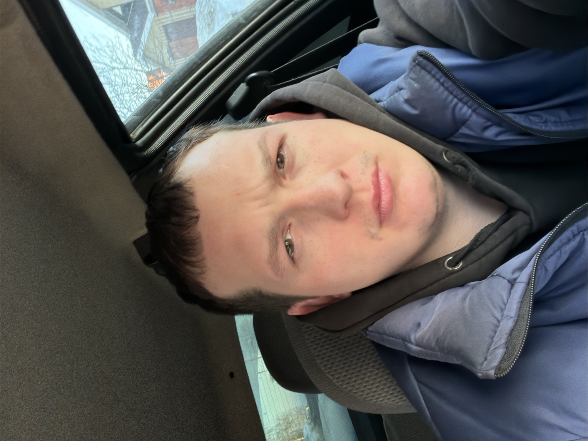 A male individual, approximately 25-35 years old, is positioned in the driver or front passenger seat of a vehicle. He has short brown hair, light skin, brown eyes, and a faint mustache, looking directly into the camera with a neutral expression. He is dressed in a dark grey hooded sweatshirt layered under a blue padded jacket, which is zipped partially. The interior of the vehicle shows a dark grey headliner and a tan/grey fabric car seat headrest behind the subject. Outside the passenger window on the right, a winter scene in Chaikovsky, Russia, is visible. Snow is present on the ground, and a bare-branched deciduous tree stands against a bright, overcast sky. In the background, a wooden house with a red exterior and a visible gable roof is discernible. The scene is illuminated by natural daylight.