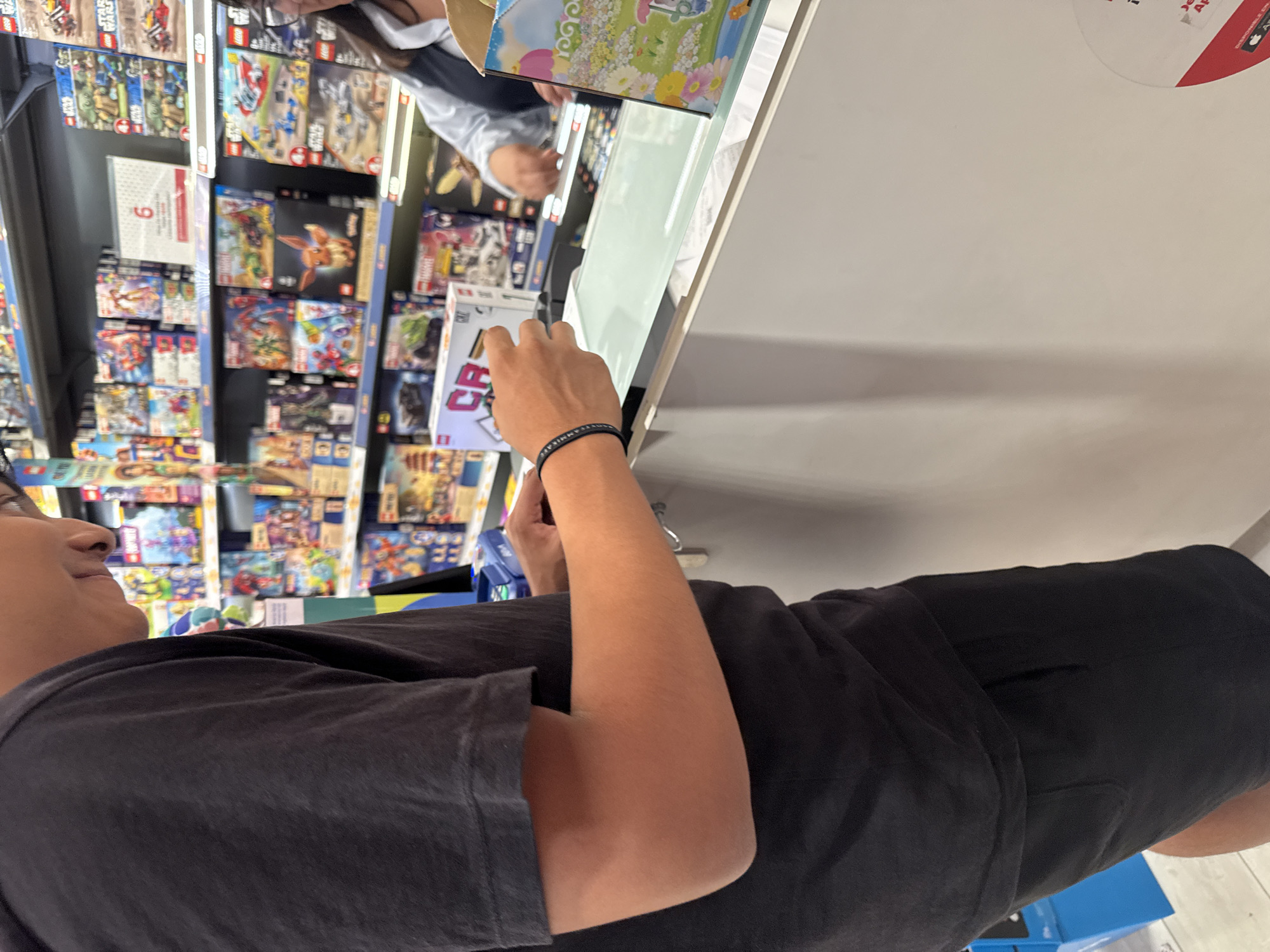 A person wearing a black t-shirt and black shorts stands in a store aisle, examining a toy on a shelf. The shelves behind them are stocked with numerous boxed toys. The location appears to be a retail store in San Bernardino Tlaxcalancingo, Mexico.