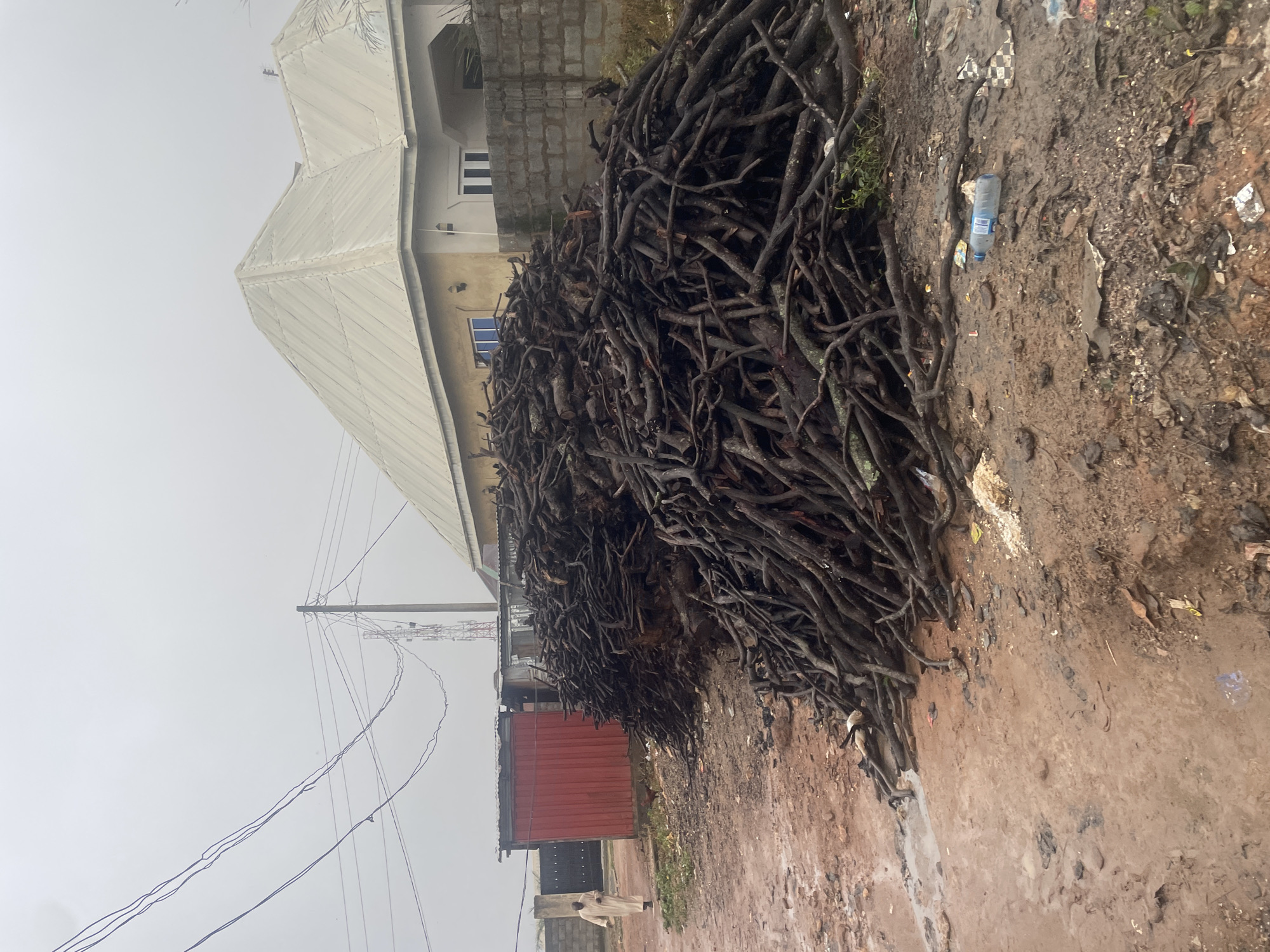 A pile of firewood is located in front of a residential building in the city of Kurunduma, Nigeria. The firewood is made of dark brown branches of varying thickness, piled haphazardly on muddy ground. To the left of the firewood, a red metal structure, possibly a kiosk or storage unit, is visible. Further left, a person dressed in a cream-colored robe walks away from the viewer on a dirt path. A grey brick wall separates the residential building from the street. The building itself is light yellow with a white, corrugated metal roof. Overhead, power lines crisscross the grey, overcast sky. The ground is a mixture of mud and debris, with a plastic water bottle visible among the scattered trash.