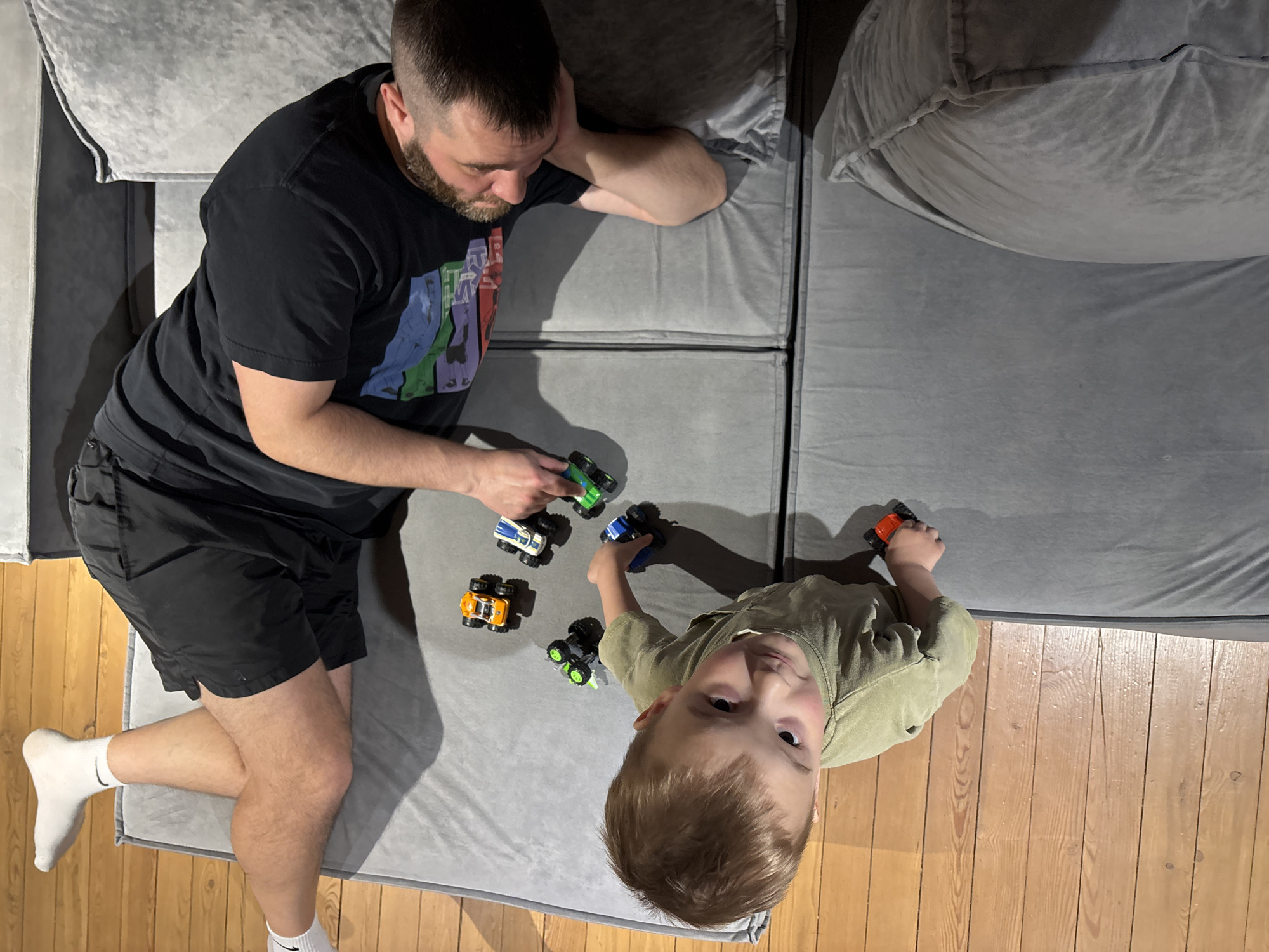 An overhead photograph captures an adult male and a young child engaged in play with toy monster trucks on a gray upholstered floor sofa or cushions in Chaikovsky, Russia. The indoor setting features wooden floorboards visible on the right and bottom edges of the frame, suggesting a home environment like a living room or play area.
The adult, a man with short brown hair and a beard, is positioned on the left side of the frame, lying on his left side with his head propped on his arm. He is wearing a black t-shirt with a colorful graphic print featuring stylized character silhouettes and fragmented, difficult-to-read text, along with black shorts and white socks. His gaze is directed downwards, focused on the toys. He holds a green and blue monster truck in his right hand.
The child, a young boy with light brown hair, occupies the lower right portion of the image. He is sitting or lying on the gray surface, looking directly up at the camera with an open, engaged expression. He wears a light olive-green long-sleeved shirt and holds an orange toy car in his right hand. Several other toy monster trucks, including yellow, blue, and green ones, are spread out on the gray surface between the two individuals.
The lighting appears to be artificial, casting soft shadows and highlighting the texture of the gray fabric and wooden floor. The overall scene depicts a calm and playful interaction between the adult and child. No specific time of day or weather is discernible from the indoor setting.