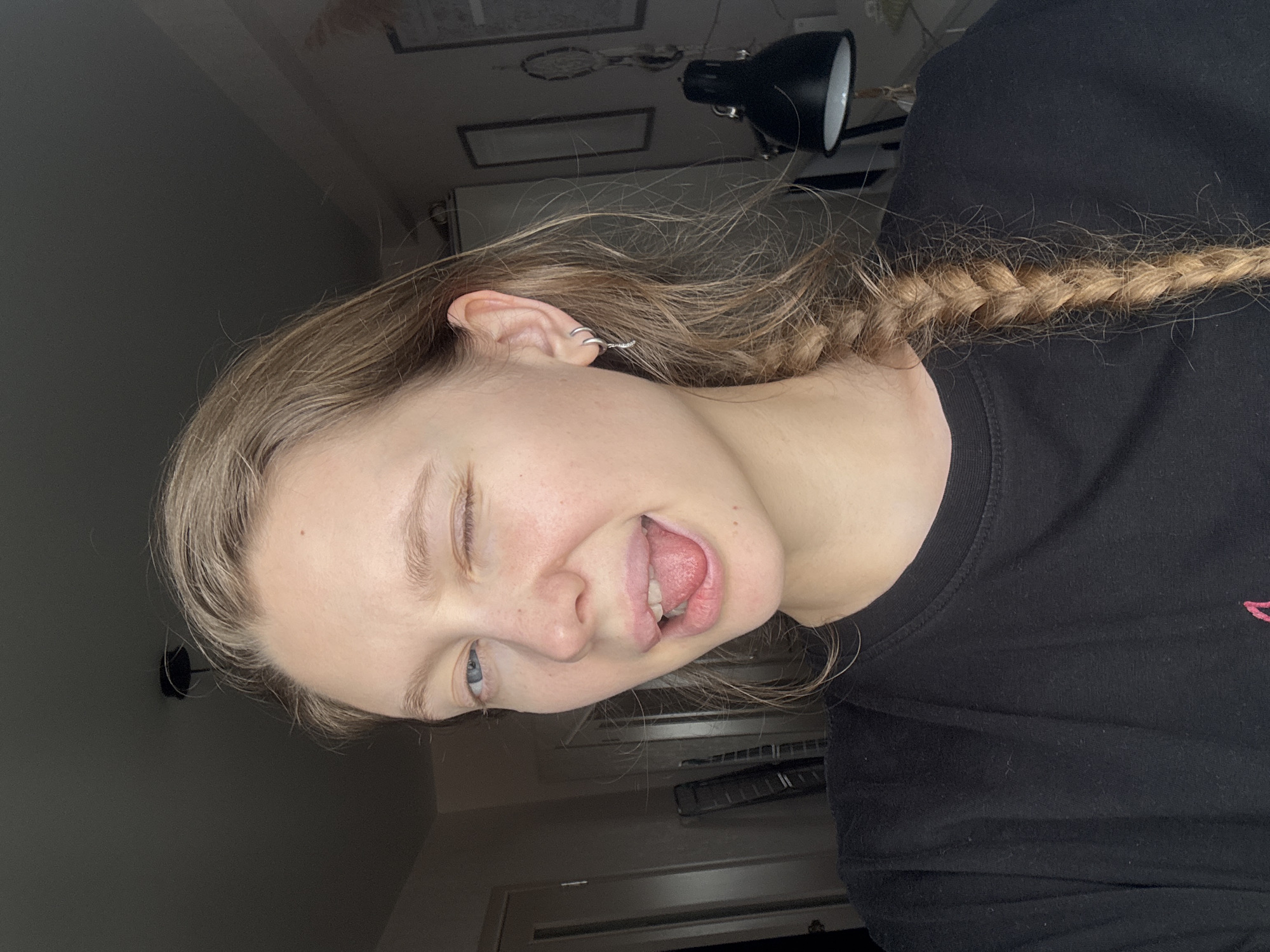The image is a portrait of a young individual, likely female, situated indoors in what appears to be a casual room setting in Izhevsk, Russia. The subject has long, light brown hair styled in a braid that extends over their right shoulder and is wearing a plain black long-sleeved top.
The person's head is tilted to the left, and they are shown with a playful expression, winking with their left eye while their tongue is slightly extended. Their eyes are blue. The left ear features multiple small, silver hoop earrings, indicating body piercings.
The background is dimly lit and consists primarily of a dark-colored wall. Visible in the upper right corner is a black desk lamp, suggesting an interior workspace or personal room. Faint outlines of framed items can also be seen on the wall behind the subject. The overall atmosphere conveyed by the subject's expression is lighthearted and cheeky. There is no visible text in the image.