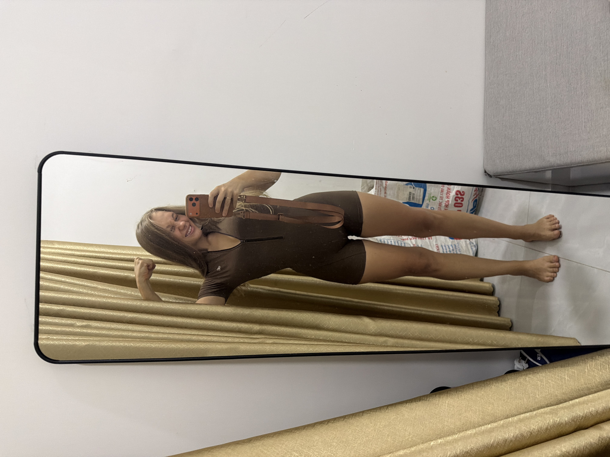 A young woman is taking a mirror selfie in Bac Nha Trang Ward, Vietnam. She is wearing a brown, short-sleeved, one-piece swimsuit with a zipper down the front and a small logo on the chest. She has a brown purse slung across her body. She is smiling and flexing her bicep. The background shows a plain white wall, a gold pleated curtain, a stack of white and blue bags with Vietnamese text, and a light gray ottoman. The floor appears to be tiled. The time of day is not discernible, and there is no indication of the weather.