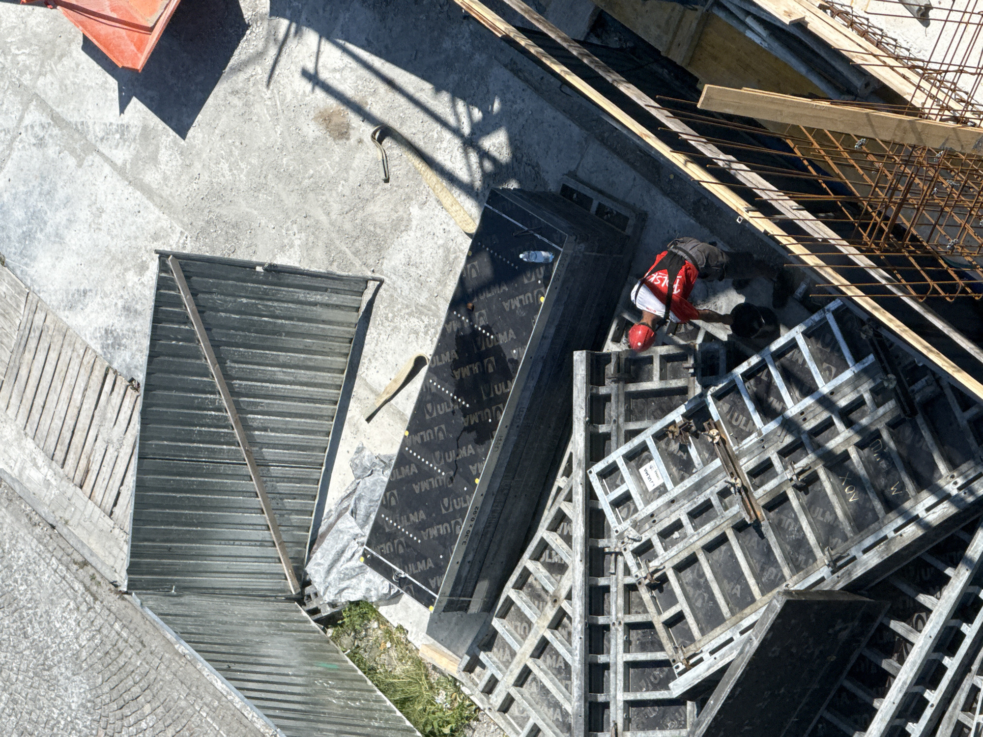 The image depicts a construction site in Gdynia, Poland. The scene is viewed from an elevated perspective, looking down onto a concrete surface where various construction materials and equipment are present. In the foreground, a construction worker wearing a red helmet and a red and white vest is visible, appearing to be engaged in a task. He is next to a black bucket and what looks like a piece of reinforcement bar.
Dominating the mid-ground are stacks of large, dark rectangular panels, possibly formwork, with "ULMA" printed on them. These are laid on top of other metallic scaffolding-like structures. To the left, a corrugated metal fence encloses a section of the site, with some green vegetation visible beyond it. Further back, a large orange bin or container casts a distinct shadow. The ground is primarily concrete, with some areas of gravel and a section of wooden planks. Shadows suggest a bright, sunny day. The overall impression is one of an active construction project in an urban environment.