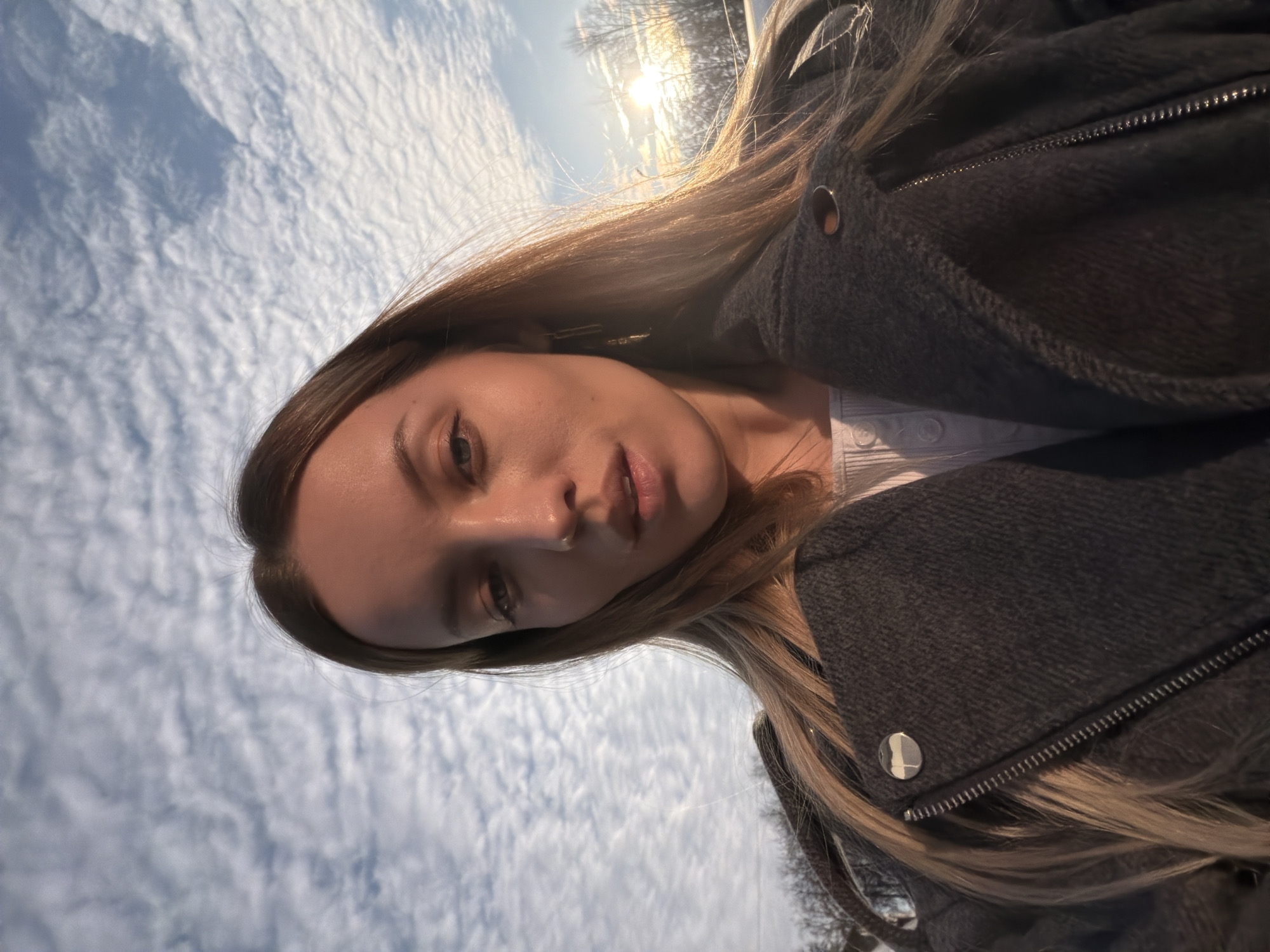 A close-up, upward-angled self-portrait captured in Klin, Russia, features a young adult female. The individual, centrally positioned, has long, light brown hair, a nose piercing on the left nostril, and wears a dark grey textured jacket with a visible zipper and a silver-colored snap button, over a light-colored, ribbed inner garment with buttons. Her gaze is directed forward, and her mouth is slightly agape. Direct sunlight from the upper right casts a bright flare and illuminates the right side of her face and hair. The background consists primarily of a blue sky densely covered with small, rippled altocumulus clouds. A sparse treeline with visible bare branches is present on the lower horizon line, particularly on the right, under the bright sun.