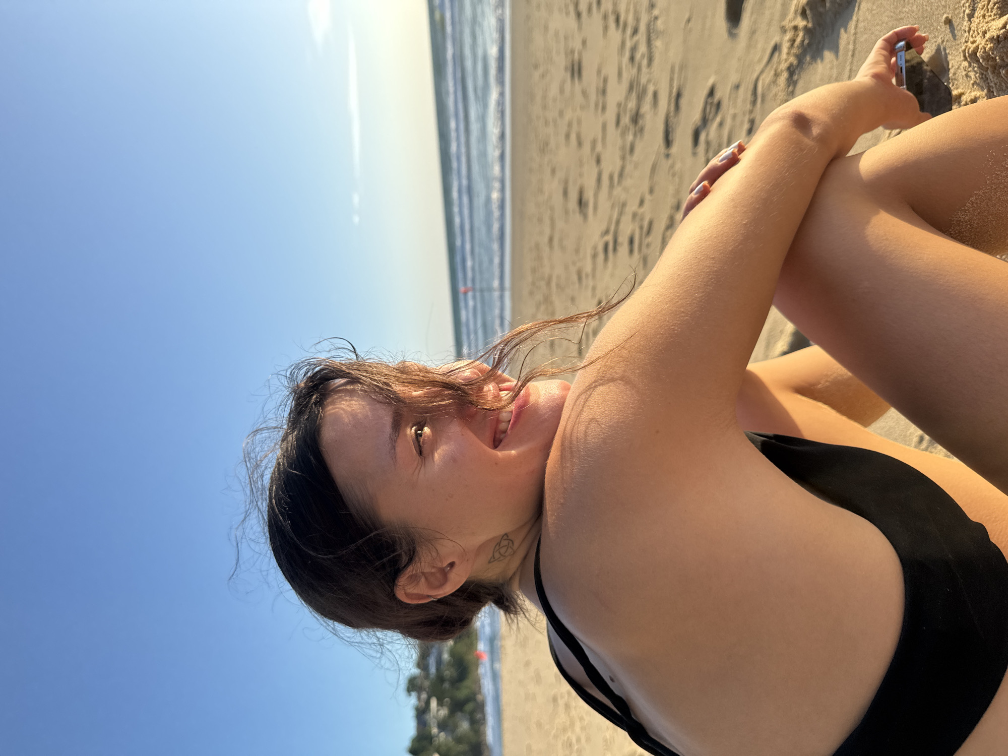 A young woman with dark hair sits on a sandy beach, turned away from the camera and looking out at the ocean. She is wearing a black bikini top. Her hair is partially obscuring her face, which is turned to her left, revealing her smiling profile. A tattoo is visible on the back of her neck. The beach is golden sand, and the ocean has gentle waves under a clear blue sky. In the distance, a red flag on a pole stands near the water's edge. The scene appears to be in Choeng Thale, Thailand.
