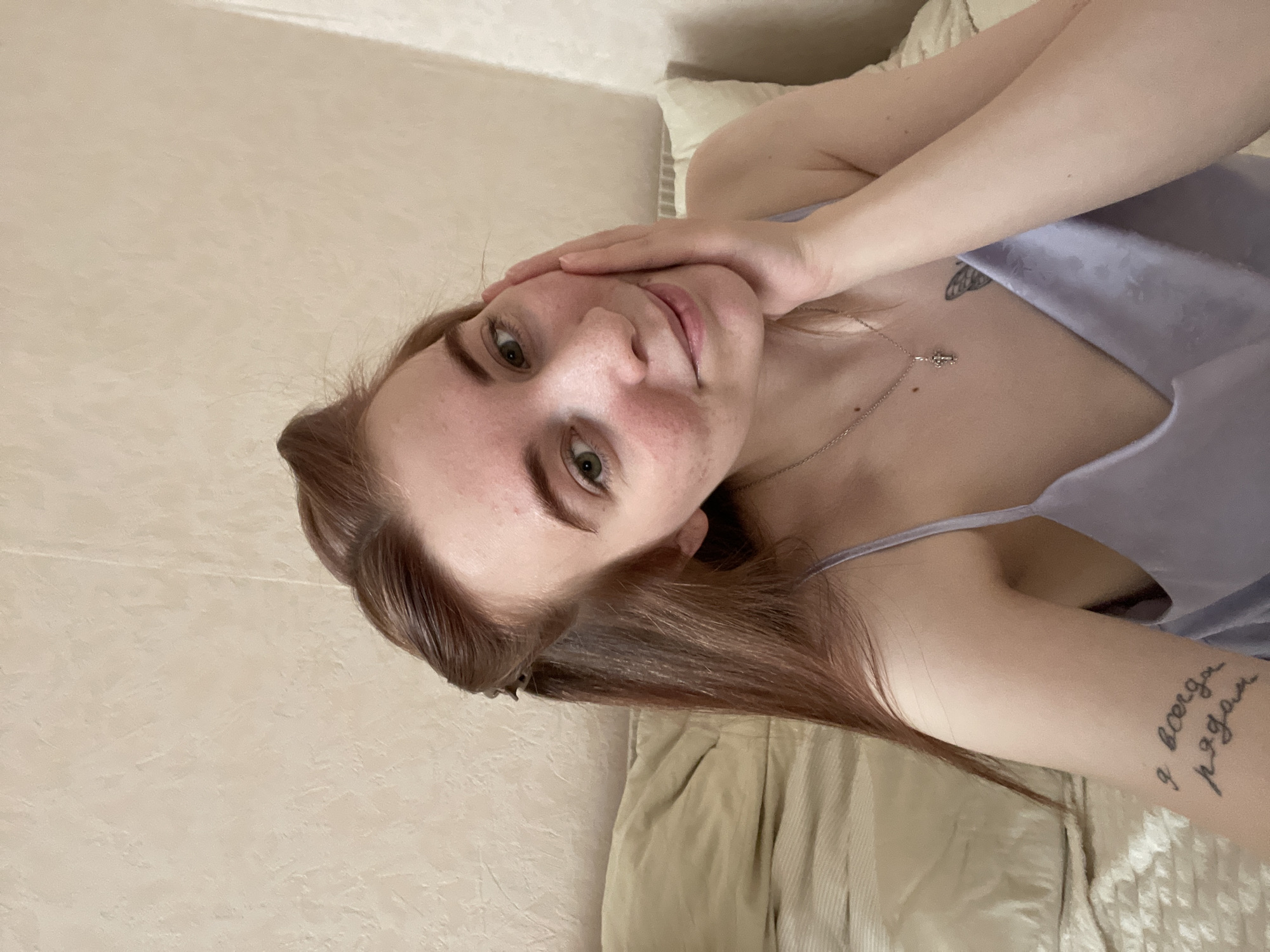A young woman with brown/auburn hair and green eyes is depicted in a close-up, tilted shot, likely a selfie. She is lying down, possibly on a bed or couch, with a light-colored, textured wall or headboard behind her. She wears a light purple spaghetti strap top or dress. Her right hand is gently placed on her left cheek and jawline, framing her face. She looks directly at the camera with a neutral to softly smiling expression.
Notable details include a silver necklace with a small charm, possibly a butterfly or cross, visible on her chest. Another small, dark tattoo, possibly a butterfly, is visible on her upper chest/collarbone area. On her left forearm, there is a prominent text tattoo in Cyrillic script that reads "Я всегда рядом," which translates from Russian to "I am always there" or "I am always near." Her skin appears fair, with some visible redness or minor blemishes on her forehead and cheeks. The lighting suggests daytime indoors.