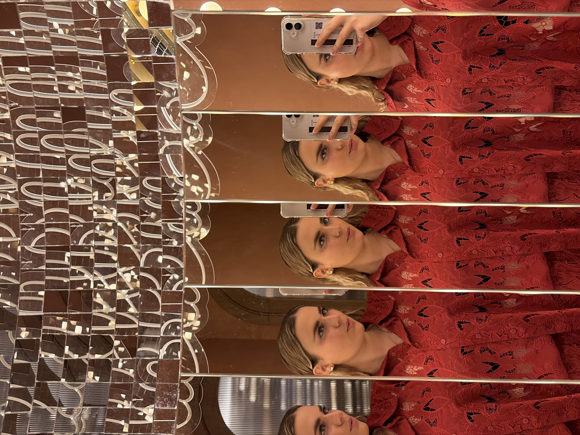 The image presents a series of reflections of a woman taking a photo of herself using a smartphone. She is wearing a red lace top. The reflections are distorted by the multi-paneled mirrored surface in front of her. To the left of the reflections, there is a mosaic-like wall decoration with repeating patterns of curved lines and mirrored pieces.
While specific architectural cues are not visible in the image, the location might be in Miami, United States of America. However, without further context or visual evidence, confirming this statement remains speculative.