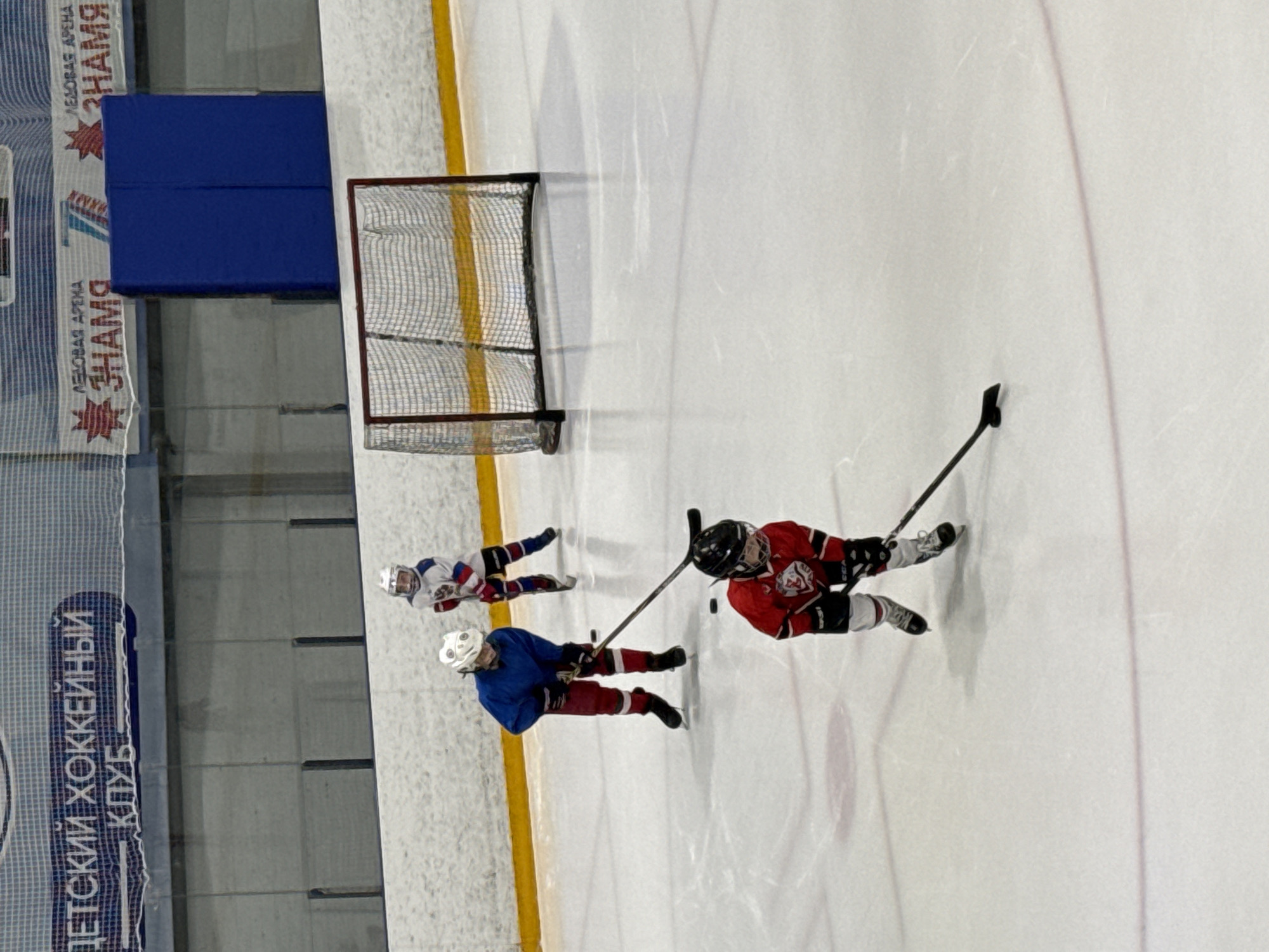 Three children, dressed in ice hockey attire, are on an ice rink. The child in the foreground, wearing a red jersey, is looking down and holding a hockey stick. To the left and slightly behind, a child in a blue jersey and red pants is also holding a hockey stick. Further back and to the right, a child in a white and red jersey is skating. A hockey net is visible in the background. The environment appears to be an indoor ice arena. Based on the text visible on the upper banner, which reads "ДЕТСКИЙ ХОККЕЙНЫЙ КЛУБ" (Children's Hockey Club), and "ЛЕДОВАЯ АРЕНА" (Ice Arena) with the logo "ЗНАМЯ" (Banner), this scene is likely taking place at the Ice Arena "Znamya" in Votkinsk, Russia.