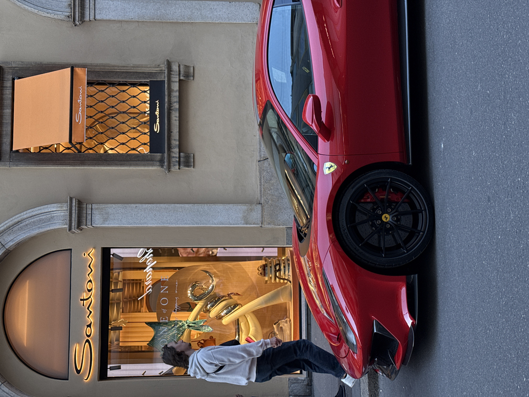 A red Ferrari is parked beside a Santoni store in Milan, Italy. A young man is standing, looking at the shop window. The Santoni shop window and awning are visible. The street is paved.