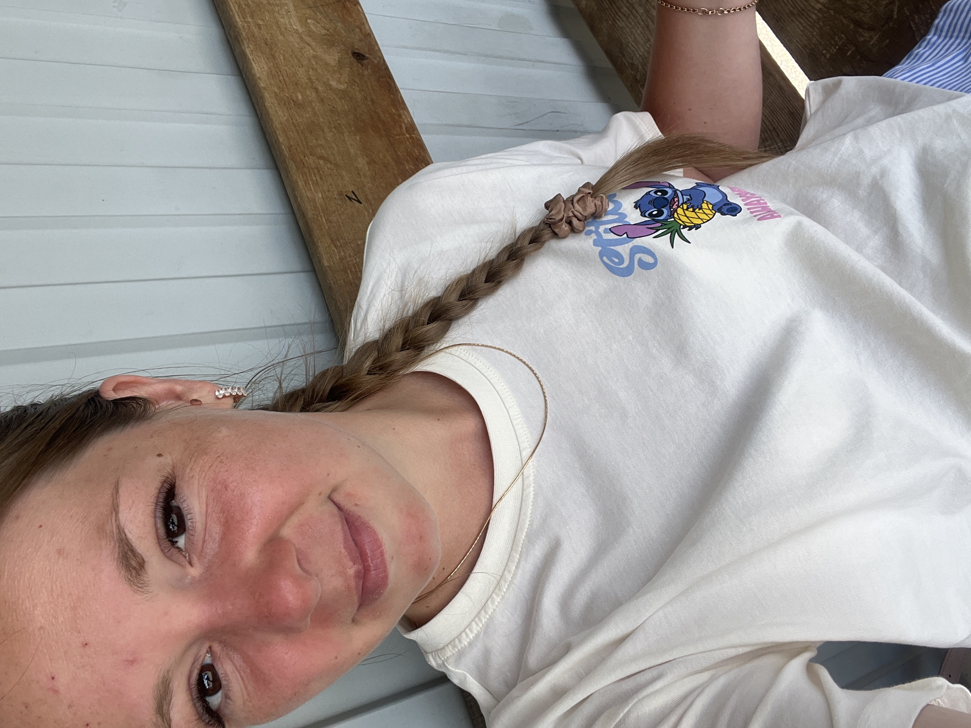 The image shows a young woman in a close-up, horizontally oriented selfie. She is wearing a light-colored t-shirt with a graphic of Stitch holding a pineapple on it, and the word "Aloha" in pink text. Her hair is braided and held with a scrunchie. She is also wearing a delicate gold necklace and a small, sparkly earring. The background appears to be an outdoor setting with a wooden plank and a light-colored, ribbed wall, suggesting a porch or deck area. The lighting indicates it is daytime, likely sunny. The woman is smiling at the camera with a relaxed and happy expression. No other individuals are visible. The image was taken in Shadrinsk, Russia.
