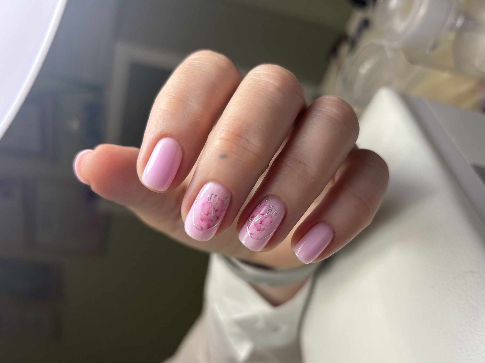 A medium shot captures a human hand with manicured nails. The nails are painted a pale pink color, with the ring and middle fingernails adorned with floral designs encircled by silver-colored lines. The hand is positioned against a backdrop consisting of a blurred light green wall, a white surface, and indistinct white objects. The city of Ферма, Russia is not discernible in this image.