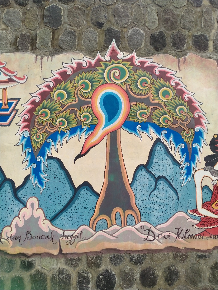 A vibrant mural in the traditional Indonesian Wayang style is painted on a light-colored wall, framed by rough-hewn grey and brown stones above and below. The central element of the mural is a highly stylized, tree-like figure, reminiscent of a *Gunungan* or *Kayon* from shadow puppet theatre. Its broad, ornate canopy is rich with patterns in green, yellow, and red hues, featuring prominent swirling motifs. A striking, elongated teardrop or flame shape forms the center of the canopy, showcasing a rainbow of colors including red, orange, yellow, and shades of blue. The tree's dark brown trunk extends downwards, with stylized roots.
Behind the tree, undulating blue mountains with a textured, stippled appearance rise. To the far left, a portion of a tiered pagoda-like structure with a red roof is visible. On the far right, a seated figure, likely a deity or character from folklore, is depicted with red and gold attire, characteristic of traditional Indonesian art. The overall scene is static and decorative, presented as a piece of public art.
Visible text is inscribed on a light brown, cloud-like element at the bottom of the mural. On the left side, partially cropped, the text reads "day Buncak Jagil." On the right side, also partially cropped, it reads "Dewi Kilosuci." The mural's intricate details and color palette suggest a cultural or mythological narrative.