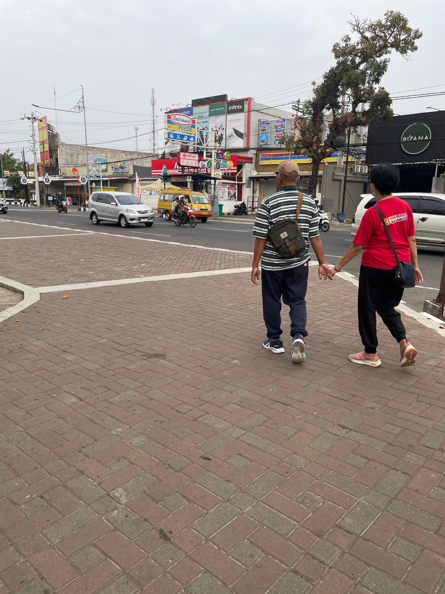 In the city of Purworejo, Indonesia, on an overcast day, an elderly couple walks hand-in-hand along a brick-paved sidewalk. The man, wearing a horizontally striped green and white t-shirt, dark pants, and a baseball cap, carries a crossbody bag. The woman, in a bright red t-shirt and black pants, has a dark bag slung over her shoulder. Behind them, a busy street features several vehicles, including a silver SUV, a yellow van, and motorcycles. Commercial buildings with prominent signage, such as "Indomaret" and "RIFANA," line the street, indicating a commercial area. The environment appears to be an urban setting with moderate traffic.