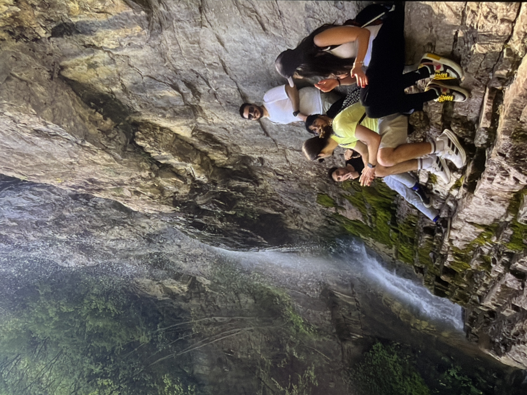 Six individuals are seated on a rocky ledge near a waterfall cascading down a cliff face. The cliff is textured with layers of stone and patches of moss. Lush green foliage covers the upper left portion of the cliff. One person, a man wearing a white t-shirt and sunglasses, stands behind the seated group, smiling at the camera. Another person, a woman with a black baseball cap, is seated on the right, facing the camera with her hands clasped. The remaining four individuals are seated on the ledge, some looking towards the waterfall and others interacting with each other. The overall environment suggests an outdoor recreational setting.