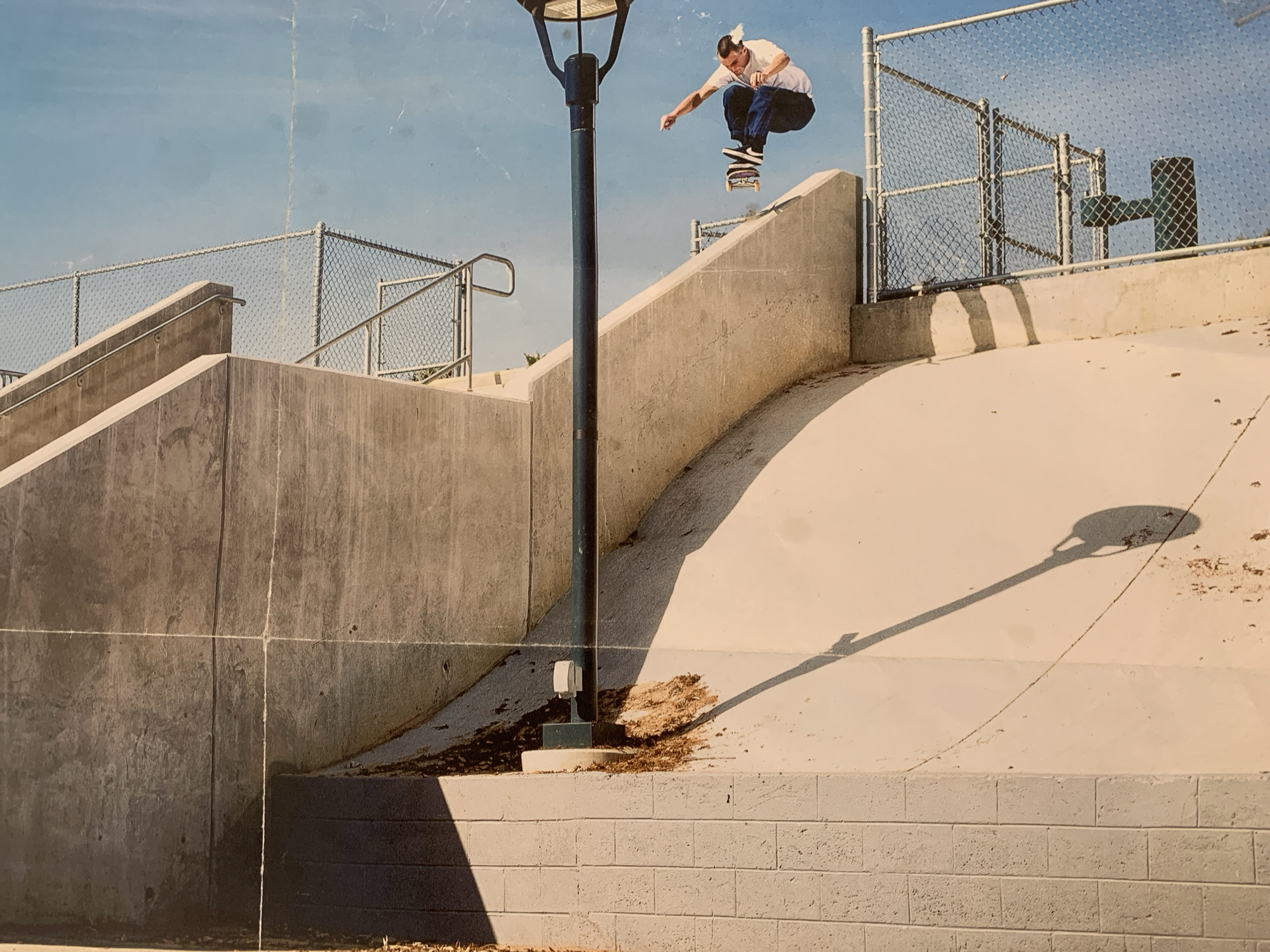 A skateboarder is captured mid-air, performing a trick on a concrete ramp. He is wearing a white t-shirt and dark pants. The skateboard is beneath his feet, indicating he is actively engaged in the sport. The environment is an outdoor urban setting with a blue sky overhead. Chain-link fences and concrete structures form the backdrop, suggesting a skate park or a similar recreational area. A tall lamp post is visible to the left of the skateboarder, casting a long shadow on the ramp. The lighting suggests it is daytime, with the sun positioned to the right. There is no visible indication of the city of Barcelona, Spain.