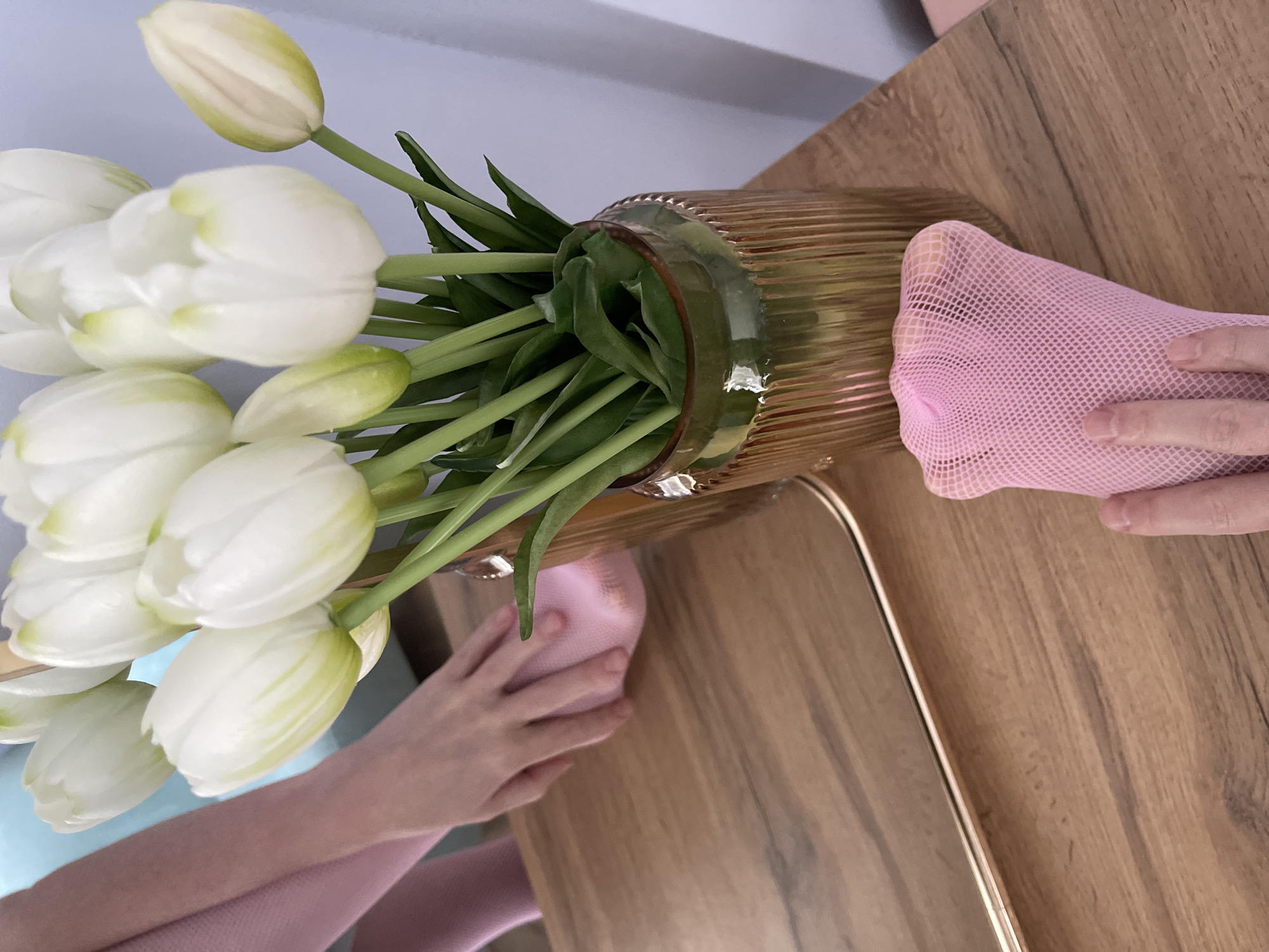 A close-up, high-angle shot shows a person's hands and legs, wearing pink fishnet tights, reaching for a bouquet of white tulips in a gold vase. The vase sits on a wooden table. A mirror on the left reflects the scene. The image is taken indoors in what appears to be a residential setting in Yablonovskiy, Russia. The lighting is soft and natural, casting subtle shadows.