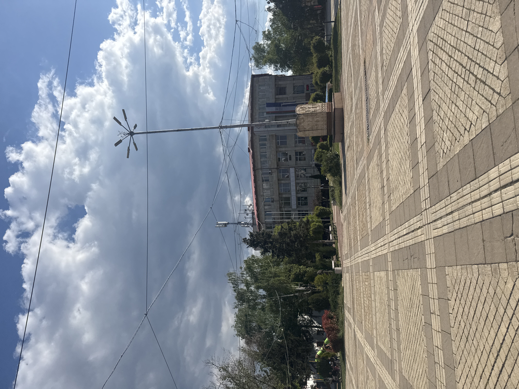 The image depicts a street scene in Elin Pelin, Bulgaria, under a partly cloudy sky. A paved area with a patterned brick design occupies the right side of the frame, leading towards a building in the distance. A tall street lamp stands to the left, intersecting power lines that crisscross the sky. Trees line the street, partially obscuring the building, which appears to be a multi-story structure with a red roof and a satellite tower. The perspective is angled, giving a sense of depth and distance in the urban landscape.