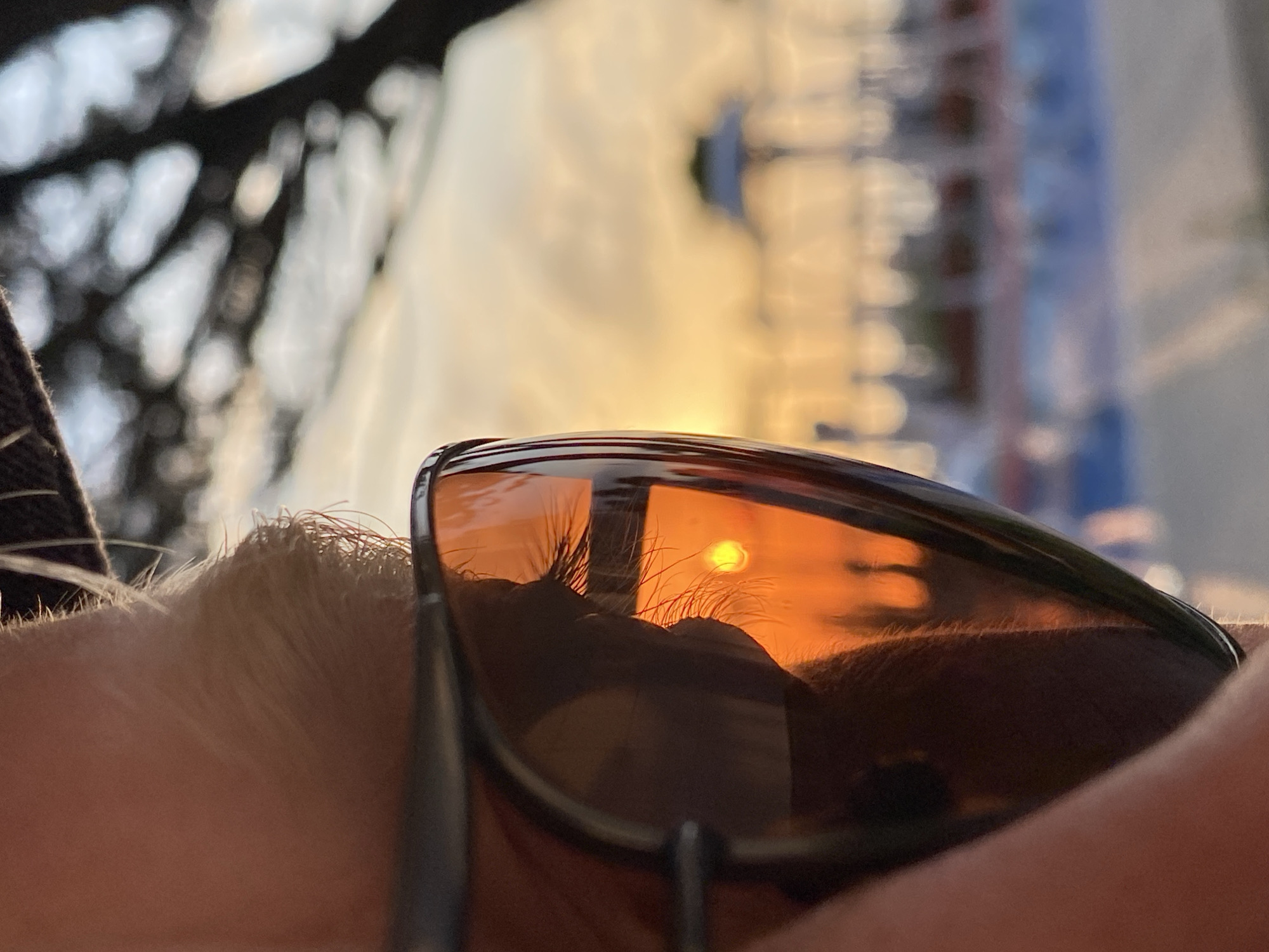 The image depicts a pair of sunglasses reflecting a sunset. The sunglasses are perched on someone's forehead, with eyelashes visible above the frames. The reflection in the lenses shows a bright sun, with potential buildings in the background, distorted by the lens curvature. The background outside the sunglasses is out of focus, but appears to show a street with building. The location of Sochi, Russia cannot be confirmed.