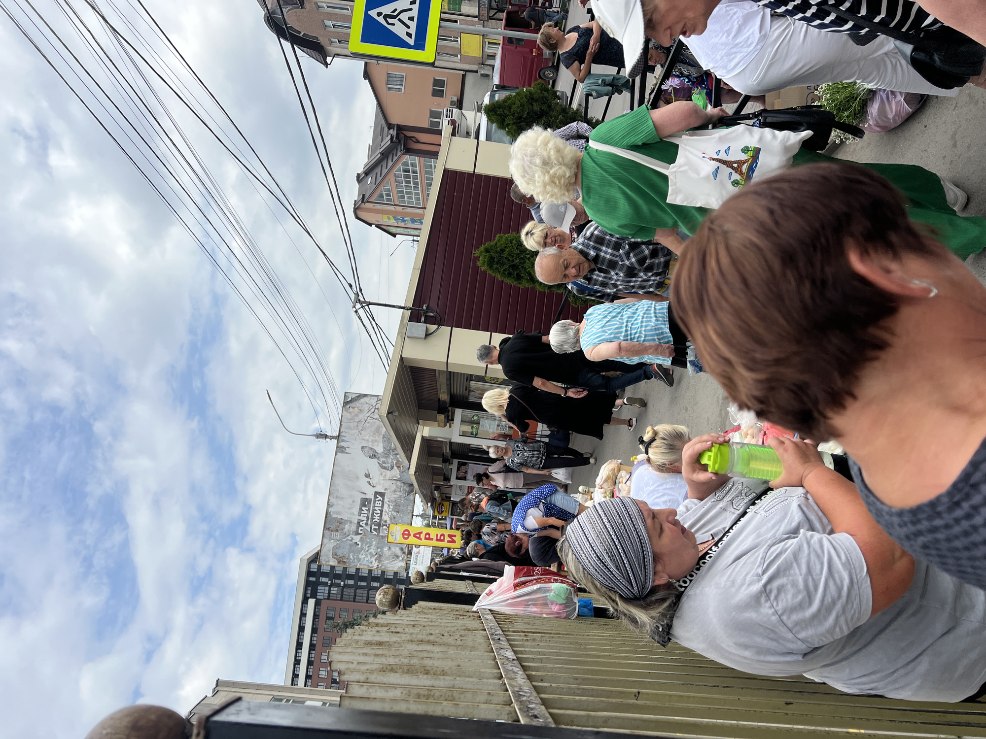 The image depicts a crowded outdoor market scene on a cloudy day, likely in Ternopil, Ukraine. A diverse group of people, predominantly adults, are navigating a narrow pathway lined with stalls and a metal fence. Many are carrying shopping bags, suggesting they are either browsing or have recently made purchases.
In the foreground, a woman with a grey headband is holding a bright green water bottle. To her right, another person's back is visible, wearing a dark patterned top. Further back, a woman in a green dress is holding a white tote bag with an image of the Eiffel Tower. Several other individuals are seen walking and interacting, including a man in a plaid shirt and an older woman with blonde, curly hair.
The environment includes various market stalls with awnings, some displaying merchandise. Behind the stalls, buildings of different architectural styles and heights are visible, including a modern apartment complex and older structures. Overhead, a network of power lines crisscrosses the sky, and a street sign with a pedestrian crossing symbol is present on the right side of the image. The overall impression is one of a bustling, everyday market atmosphere.