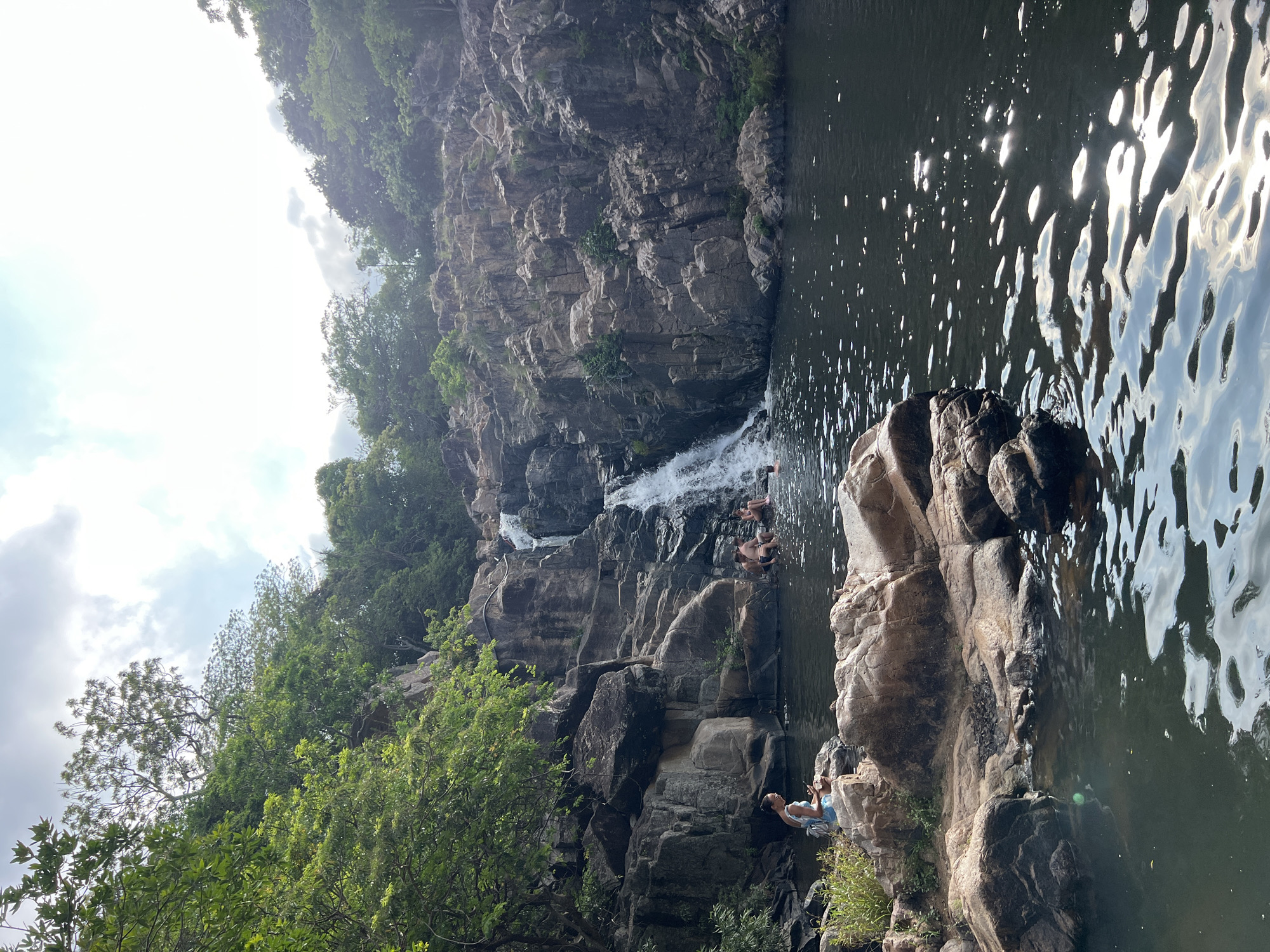 This image depicts a natural waterfall and swimming hole in Vĩnh Hải, Vietnam, under a bright, partially cloudy sky. A multi-tiered waterfall cascades down rugged, dark grey and brown rock formations on the right side of the frame, emptying into a large, dark pool of water. The pool's surface reflects shimmering sunlight, particularly in the foreground and midground. Dense green trees and lush foliage cover the rocky terrain, especially prominent on the left side and cresting the top of the rock formations surrounding the waterfall.
In the left foreground, a woman with dark hair, wearing a light blue or teal outfit, sits on a flat, light-colored rock at the edge of the pool, looking down at a small handheld device. A light-colored backpack rests beside her. In the midground, closer to the waterfall, at least four individuals are present. Two men are seated on rocks at the water's edge, partially submerged, while two other individuals are partially in the water, one near the base of the waterfall and another further to the right. The overall environment is a natural, rocky landscape dominated by water and abundant vegetation.