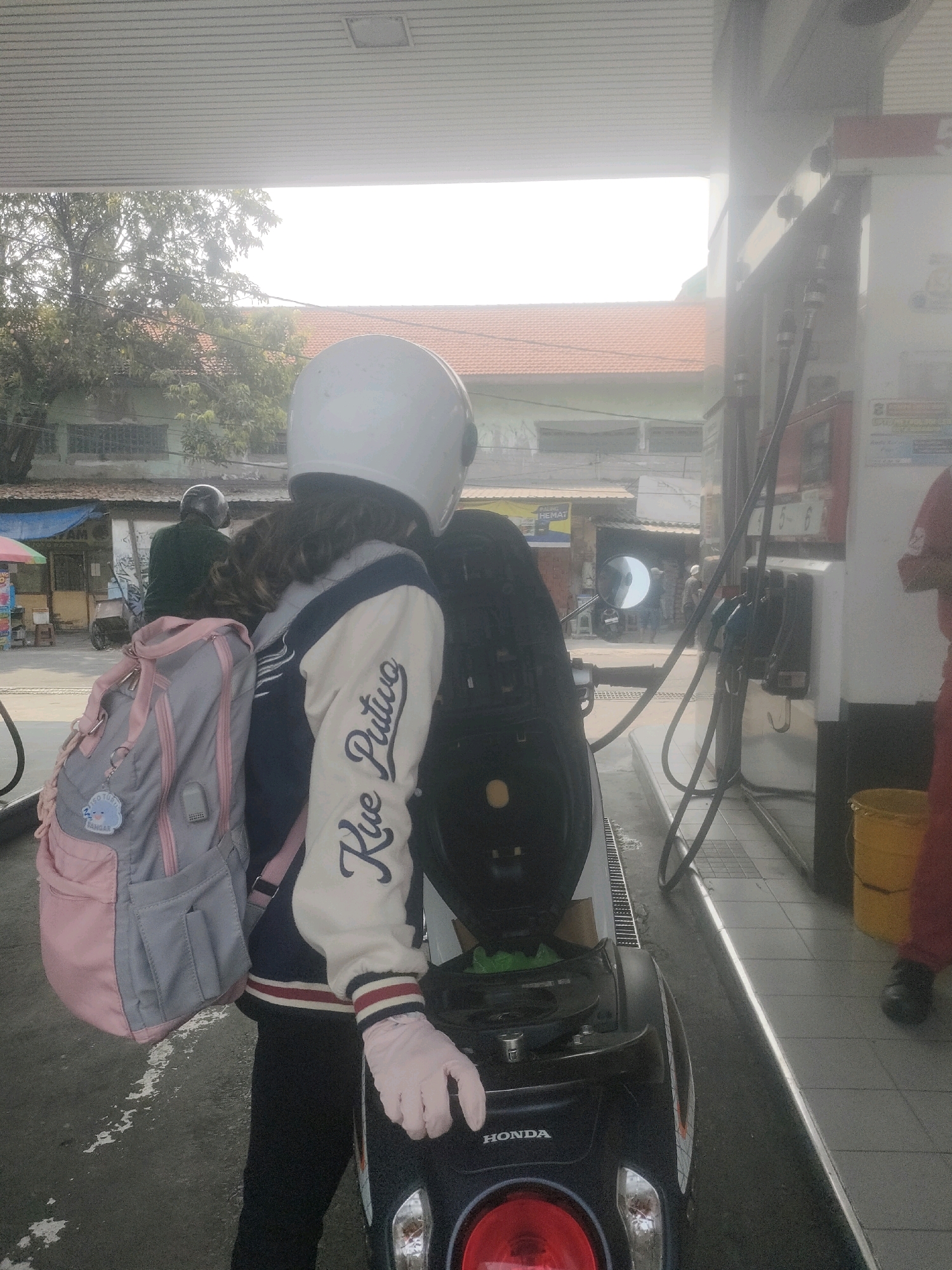 A person wearing a white helmet and a white and blue bomber jacket is standing next to a black scooter at a gas station in Surabaya, Indonesia. The person is also wearing light pink gloves and a grey and pink backpack. The scooter is open, revealing a green object inside. The gas station has a red and white sign and yellow buckets. In the background, there are buildings and trees. The sky is bright and overcast.