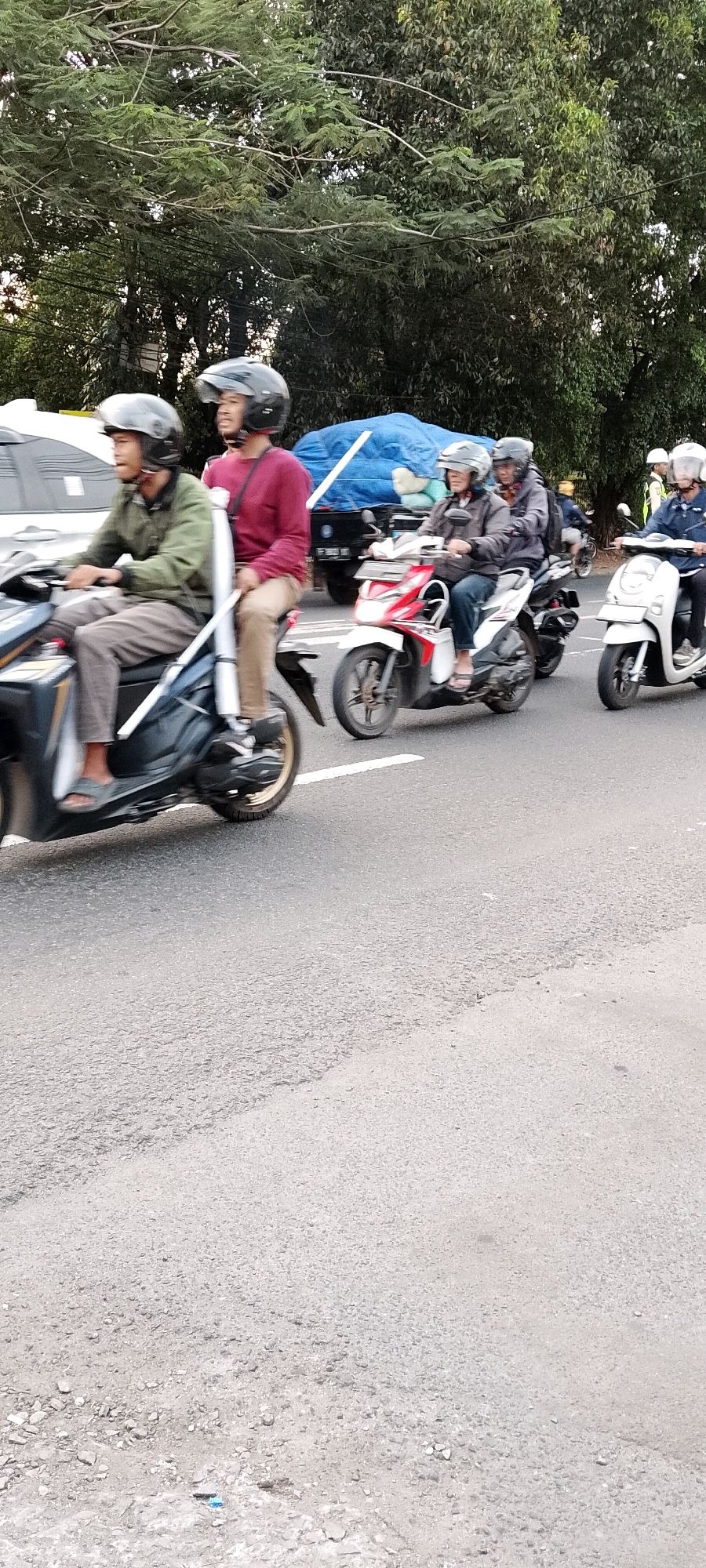 An eye-level medium shot captures a group of individuals riding motorcycles in what appears to be a street in Depok, Indonesia. The scene is dominated by the road and the vehicles, with lush green trees lining the background. In the foreground, two men are riding a motorcycle. The driver, wearing a green jacket and a black helmet, is looking to his left. The passenger, in a maroon shirt and black helmet, is looking straight ahead. Behind them, another motorcycle with two occupants is visible, a driver in a dark jacket and helmet and a passenger in a grey jacket and helmet. Further back, a white scooter with a single rider in a blue jacket and helmet is also on the road. A truck with a blue tarp-covered load is parked on the side of the road. The overall impression is one of daily urban commute, with motion blur indicating movement.