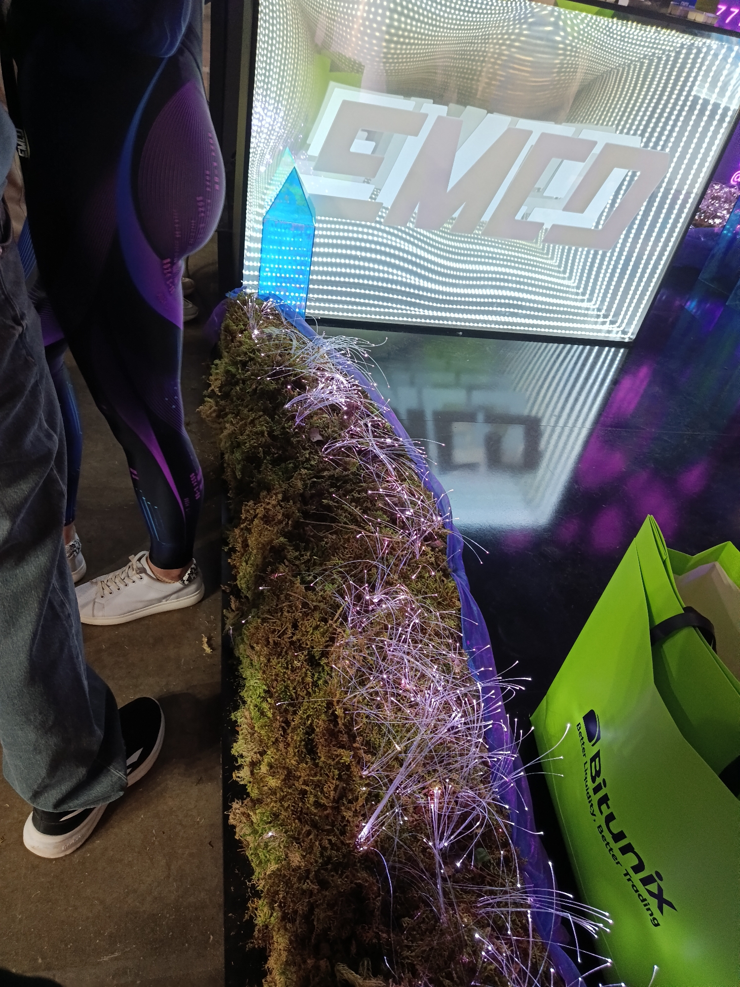 The image depicts a scene with several elements, most likely indoors. To the left, there are parts of two individuals: one wearing a dark-colored outfit with purple accents and white shoes, and another in jeans and black sneakers with white soles. In the center, there's a display of moss or greenery decorated with numerous fiber optic lights that resemble glowing white hairs. Behind the greenery, there's a light-up display with the word "GMCO" in a modern font, creating a mirror or infinity effect with the repeating word. To the right of the greenery is a green tote bag with the word "Bitunix" and the phrase "Better Liquidity, Better Trading" printed on it. Based on the available details, the location being in Khimki, Russia, cannot be confirmed.