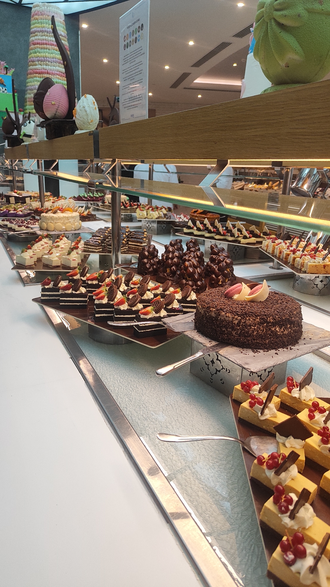 The scene depicts a dessert buffet line inside an establishment, likely a hotel or resort, in Alanya, Turkey. The foreground shows a white counter surface and a clear glass display barrier. Behind this barrier, an extensive array of desserts is presented on various elevated stands and trays.
Prominent items include a large, round chocolate cake covered in dark chocolate shavings, garnished with two pink and white confectionary pieces. To its left, multiple individual rectangular chocolate cakes are topped with half strawberries and circular chocolate biscuits. To the right, numerous individual rectangular yellow cakes are visible, each adorned with white cream, red currants, and a piece of dark chocolate. Silver serving spatulas are positioned near several dessert sections. Further back, clusters of dark chocolate-coated items are arranged, alongside a round white cake decorated with orange slices and white chocolate circles.
In the upper left background, a tall, multi-colored, tiered decorative cone stands next to a pink and a white decorative egg. A white informational sign displaying "FOOD ALLERGEN ICONS" with rows of circular symbols is visible behind a horizontal wooden beam that runs above the dessert display. The background is blurred, showing an indoor ceiling with recessed lighting and indistinct figures, possibly staff in white uniforms.