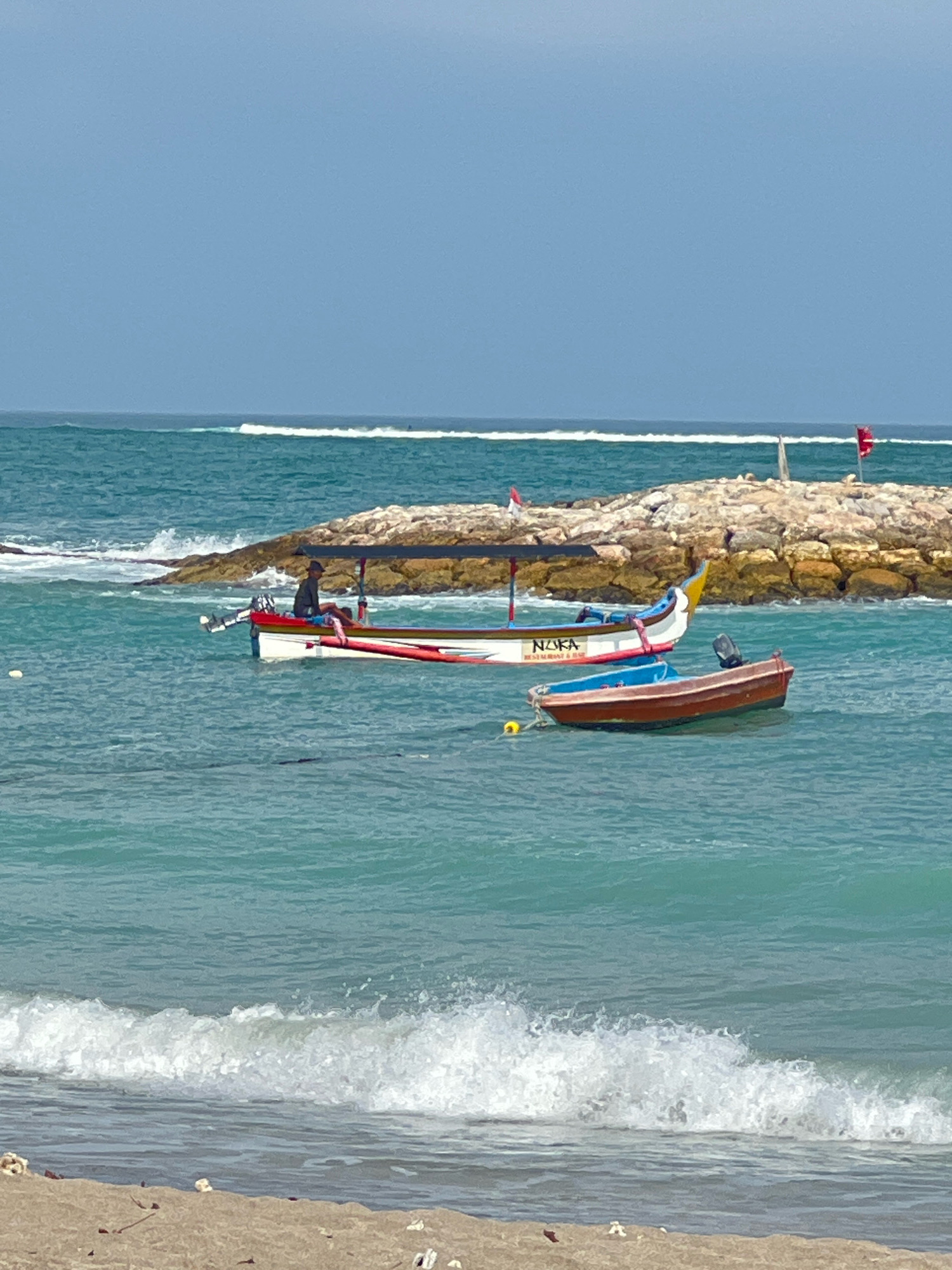 The image depicts a serene beach scene in Kuta, Indonesia. The turquoise water of the ocean laps gently at the sandy shore in the foreground. In the middle ground, two boats are anchored offshore. The larger boat, a traditional Indonesian outrigger with a canopy, is painted white with red trim and the word "NUGA" emblazoned on its side. A person wearing a hat and a dark shirt sits in the boat, seemingly relaxed. Behind it, a smaller, reddish-brown boat with a blue interior floats on the water. Further in the background, a breakwater constructed of large stones juts out into the sea, with a small red flag planted on it. The sky above is a clear, pale blue, indicating fair weather. The overall impression is one of tranquility and natural beauty.