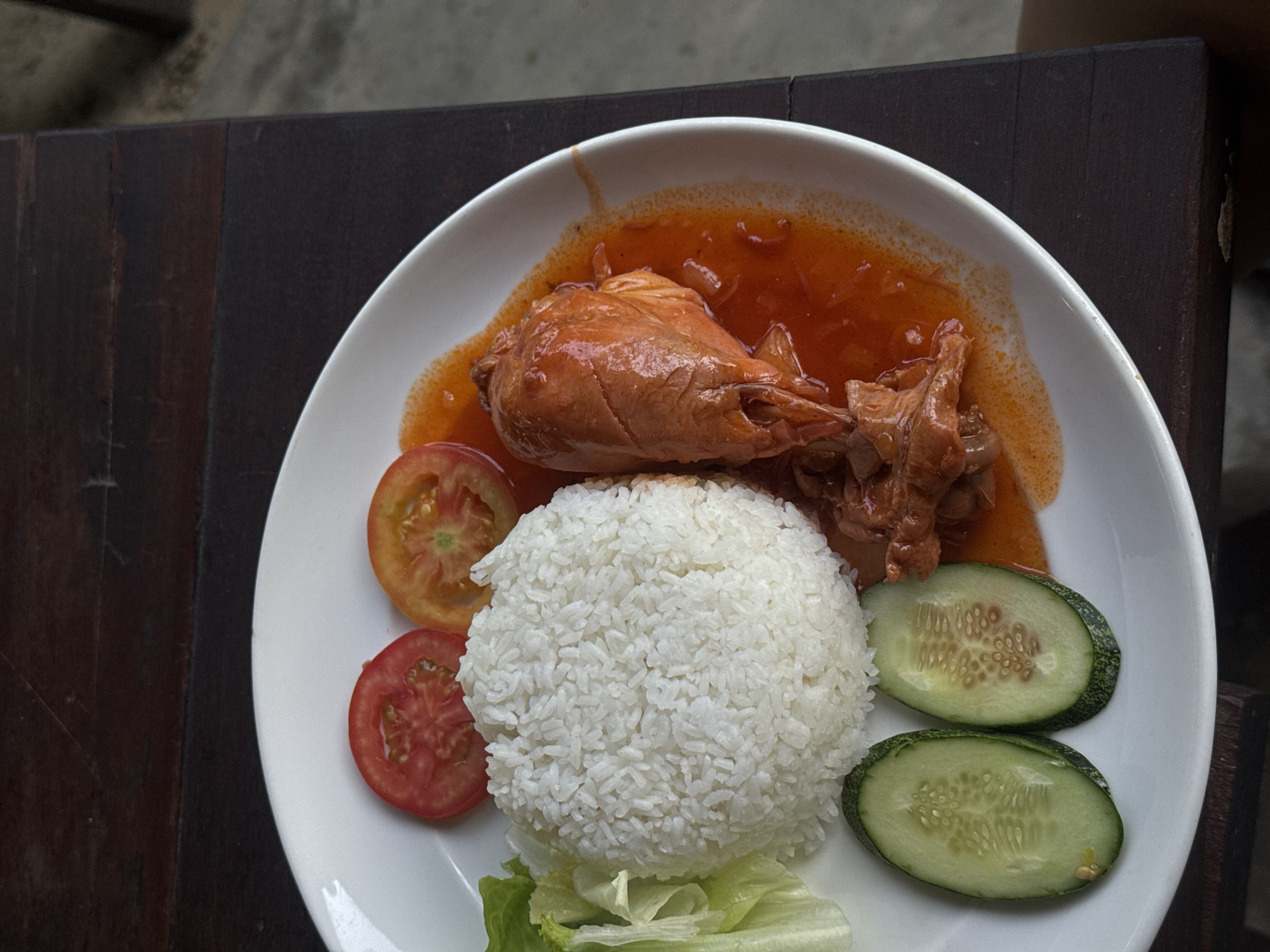 A plate of food is presented on a dark brown wooden surface. The dish consists of a portion of white rice molded into a hemispherical shape, accompanied by a piece of chicken, likely a drumstick, glazed with a red-orange sauce. Two slices of tomato and two slices of cucumber are arranged around the rice. Some lettuce is positioned near the bottom of the plate. The location is unidentified, and no people are visible in the image.
