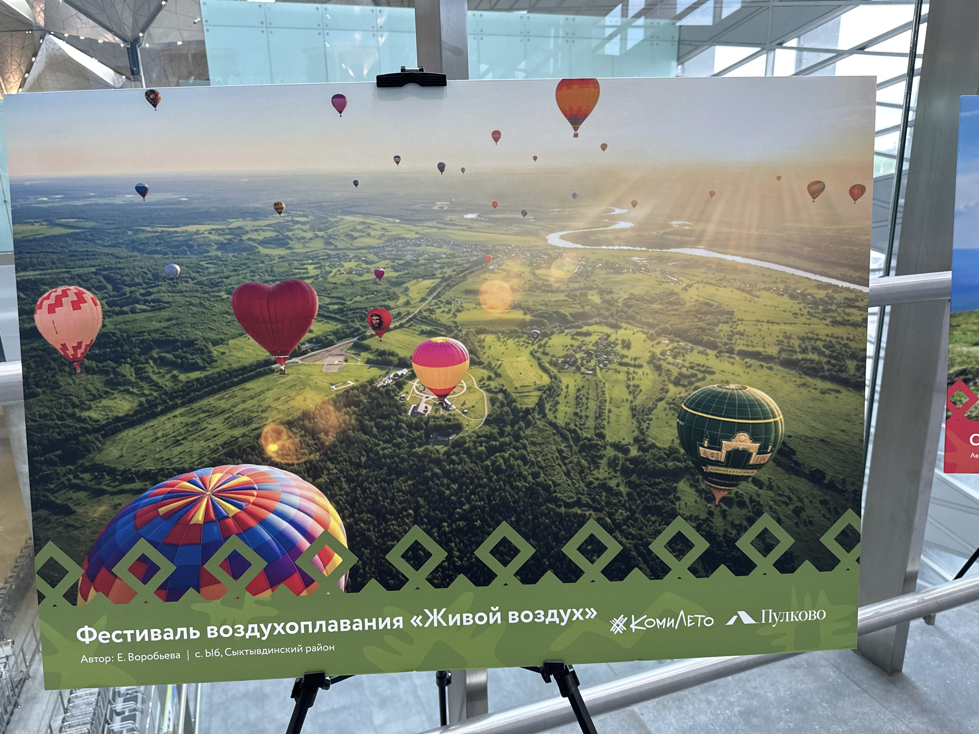 The image shows a poster on an easel, likely indoors, featuring a photograph of many hot air balloons floating over a green, landscape with a river visible in the distance. The poster's text translates to "Hot Air Balloon Festival 'Living Air'" and the author is listed as E. Vorobieva from the Syktyvdinsky region. Logos for "Komileto" and "Pulkovo" are also present. The picture on the poster is taken in Saint Petersburg, Russia.