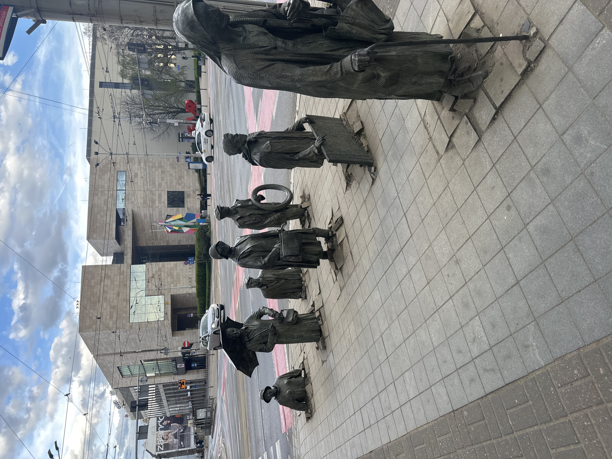This image shows a sidewalk in Wrocław, Poland, featuring the "Passage" monument. The artwork consists of bronze statues of individuals emerging from the pavement. The statues are spaced apart, depicting a procession-like formation. In the background is a multi-story building with a large glass facade and a stone exterior. On the street, red and white crosswalk markings are visible, along with street signs and overhead tram lines. A white car is parked nearby. The sky above is partly cloudy. The sidewalk itself is paved with square tiles.
