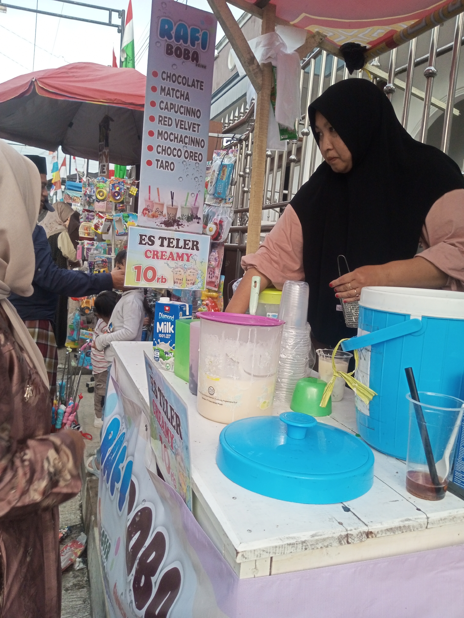 The image captures a street-side vendor stall in Pandansari, Indonesia, selling boba drinks and "Es Teler Creamy." A woman wearing a black hijab and a pinkish top is serving customers. To her left, a child in a grey sweater stands near a display of toys. Behind the vendor, a red umbrella is open, and various products are displayed, including a sign for "Rafi Boba Drink" listing flavors like Chocolate, Matcha, Capucinno, Red Velvet, Mochacinno, Choco Oreo, and Taro. Another sign advertises "Es Teler Creamy" for 10,000 Indonesian Rupiah. A blue cooler, plastic cups, and containers of ingredients are visible on the counter. The ground is paved, with some litter and a few colorful items scattered around. The scene appears to be set during daylight hours.
