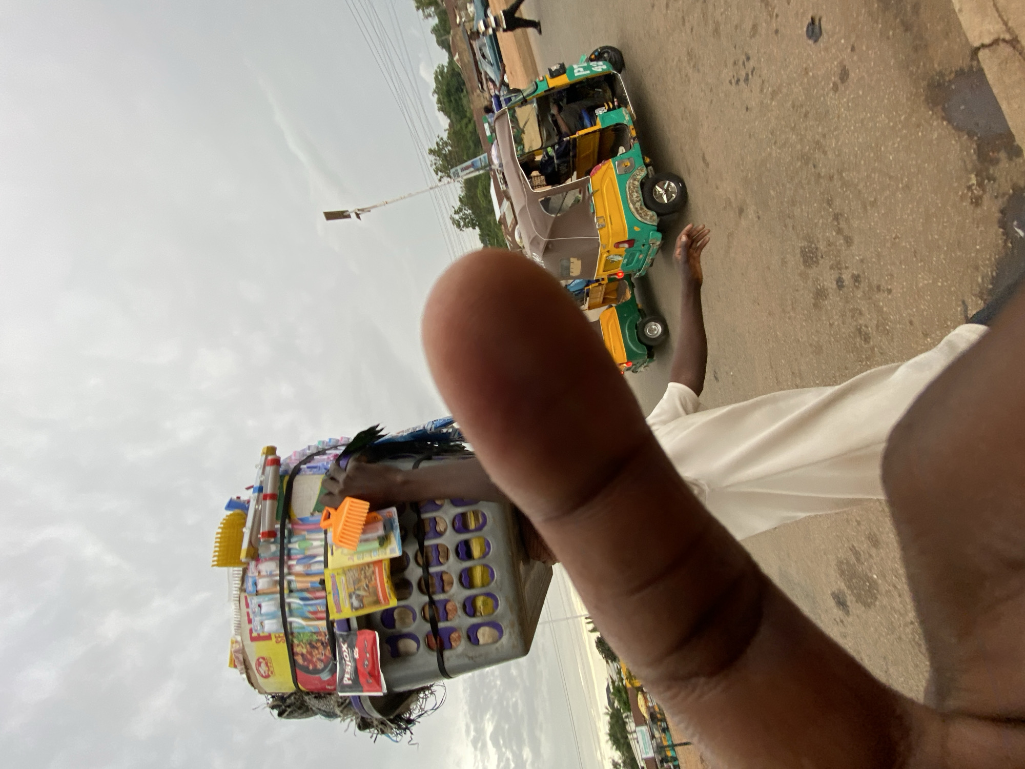 The image, taken in Bukuru, Nigeria, shows a man standing on a paved street, likely attempting to sell his wares. The items, including toothbrushes, insecticide, instant noodles, and hair combs, are stacked in a plastic basket which the man holds with his left hand. In the background, a yellow and green auto-rickshaw (tuk-tuk) is parked on the side of the road. Other people are visible walking further up the road. The sky is overcast. A portion of the photographer's finger is prominently displayed in the foreground, obstructing the view. The image is slightly tilted counterclockwise.