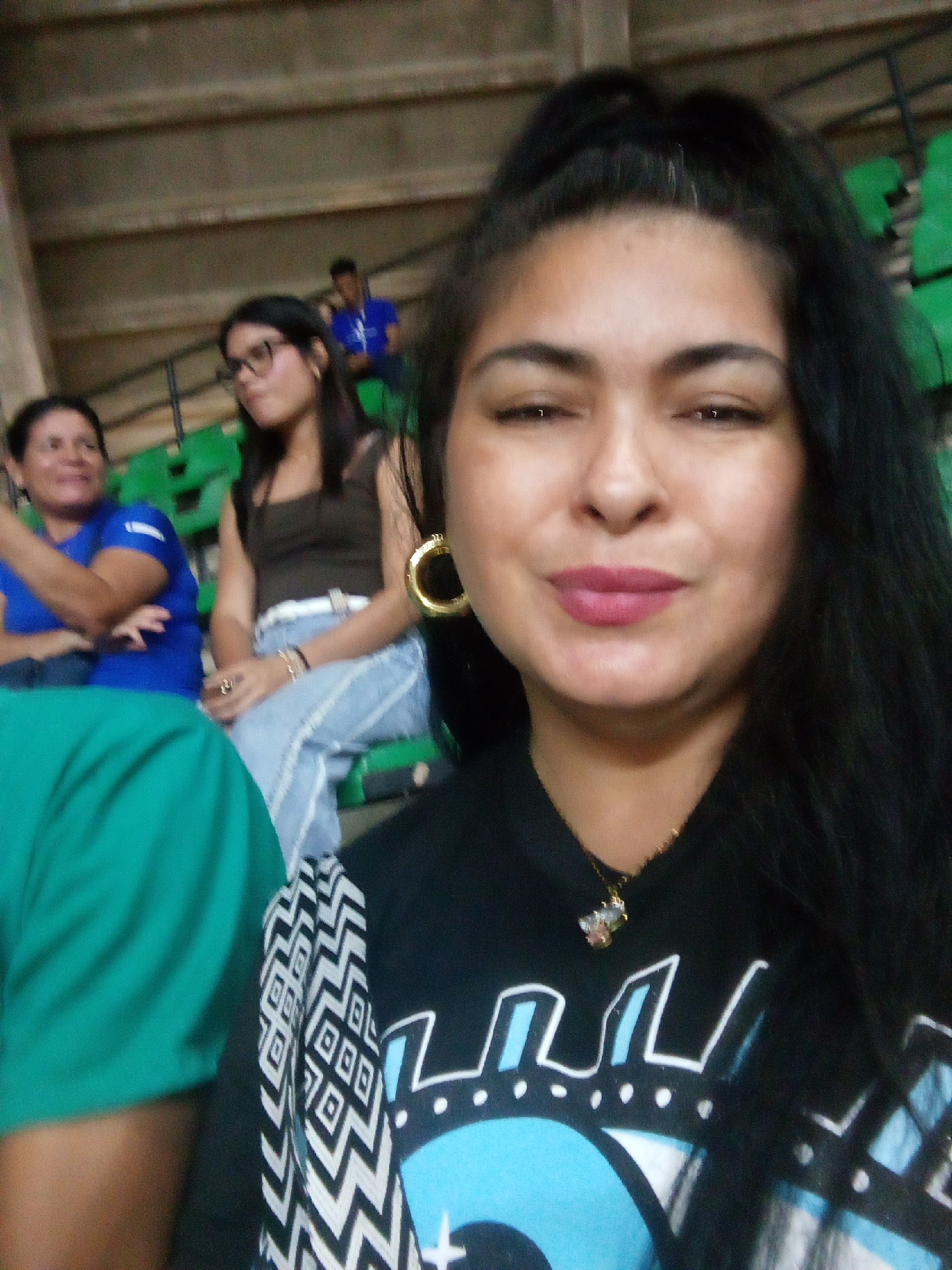 The image shows a woman in the foreground, wearing a black shirt with a blue and white graphic design and a gold necklace. She has dark hair and is wearing gold hoop earrings. Behind her, there are stadium-style seats, occupied by several other people. One woman is wearing a blue shirt, and another is wearing a brown tank top and jeans. A man in a blue shirt is also visible in the background. The location appears to be a stadium or arena setting. The city is unknown.