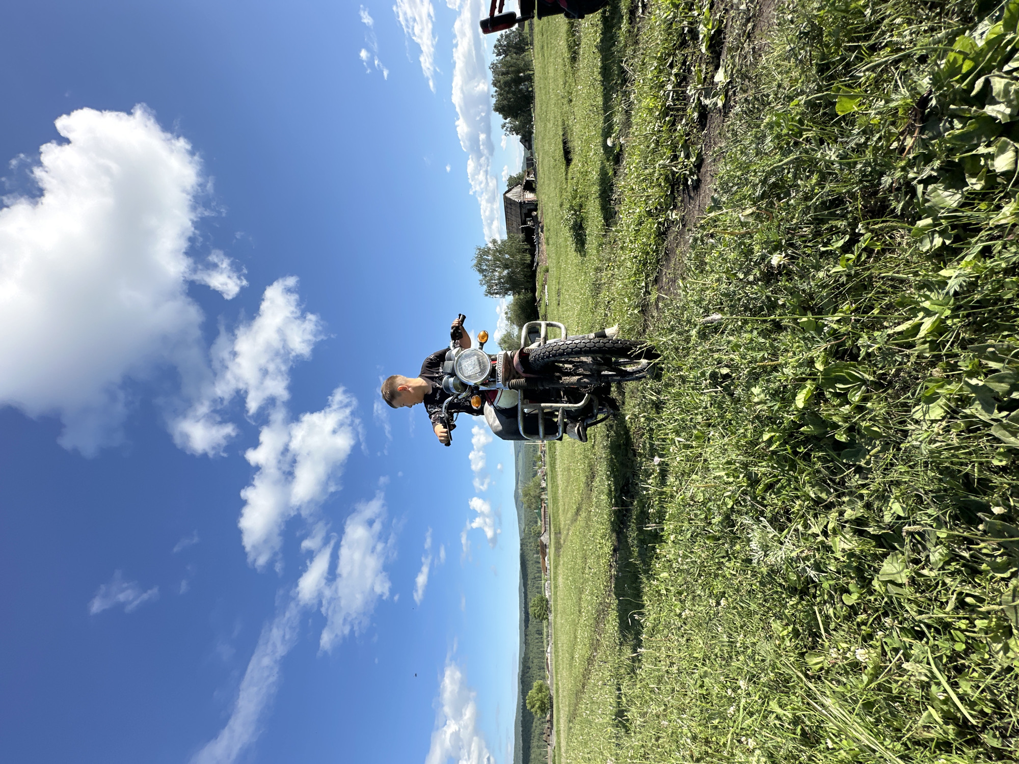 In a rural landscape under a bright blue sky with scattered clouds, a young person sits on a red motorcycle in a grassy field. The person is wearing a black shirt with a white graphic design and appears to be looking forward. The motorcycle has a circular headlight and a metal rack on the front. The field is covered in lush green grass with some white wildflowers and a dirt path running through it. In the background, there are trees, some rural buildings, and rolling hills. The image captures a moment of leisure and exploration in a natural setting.
