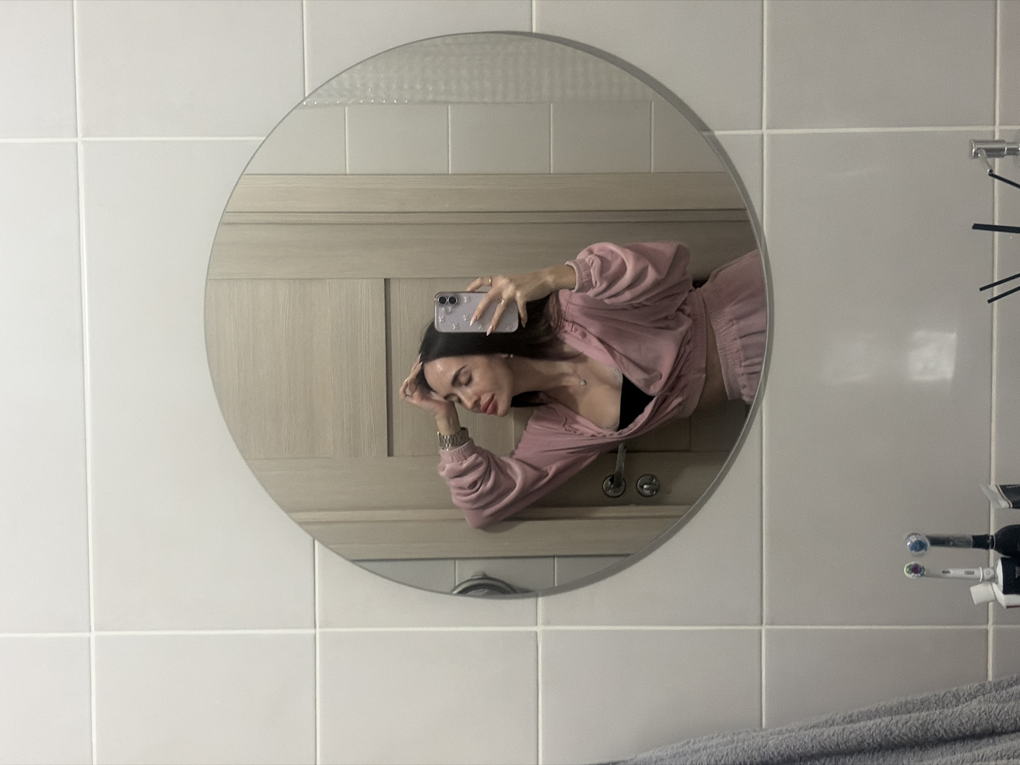 A woman in a pink tracksuit takes a selfie in a round mirror in a bathroom. The bathroom has white tiled walls and the mirror reflects a door with horizontal panels. Toothbrushes are visible to the right of the mirror. The location cannot be determined to be in Saint Petersburg, Russia.