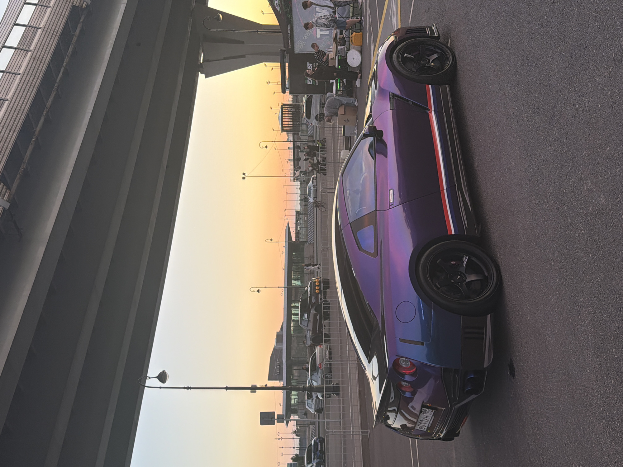 The image depicts an outdoor scene under a large overpass, with a purple Nissan GT-R car prominently featured in the foreground. The car has a unique iridescent paint job that shifts between shades of purple and blue. It has black rims and racing stripes along the side, consisting of red, white, and blue lines. The car's license plate is visible, reading "B011XH 198".
In the background, there is a street with several other cars, including a dark-colored pickup truck and a silver sedan. A group of people are gathered near a tent or booth with signage that reads "EXUST AUTOMOTIVE APPAREL". The sky shows a gradient of colors, transitioning from a bright yellow-orange near the horizon to a lighter blue, indicating it is either sunrise or sunset. There are streetlights and a distinctive architectural structure of the overpass overhead, casting shadows on the scene. The ground is paved asphalt.