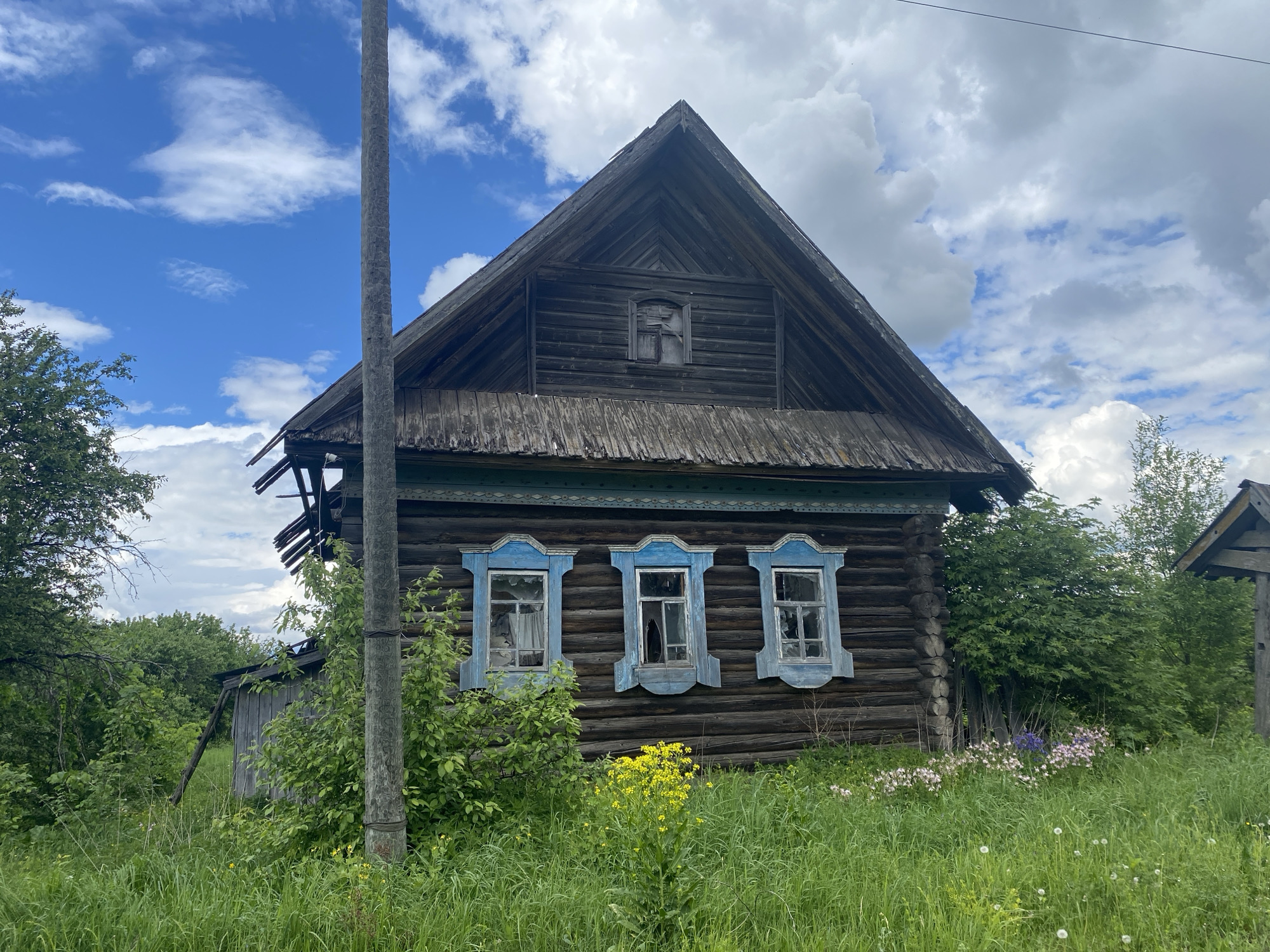 The image shows a dilapidated wooden house in Puzhmes'-Tuklya, Russia. The house has a steeply pitched roof and three windows with blue painted frames on the front facade. The windows appear broken or boarded up. The wood of the house is dark and weathered. The house is surrounded by tall green grass and various plants, indicating a state of neglect. A utility pole stands near the house, partly obscured by vegetation. The sky is partially cloudy with patches of blue visible. There are no people visible in the image.