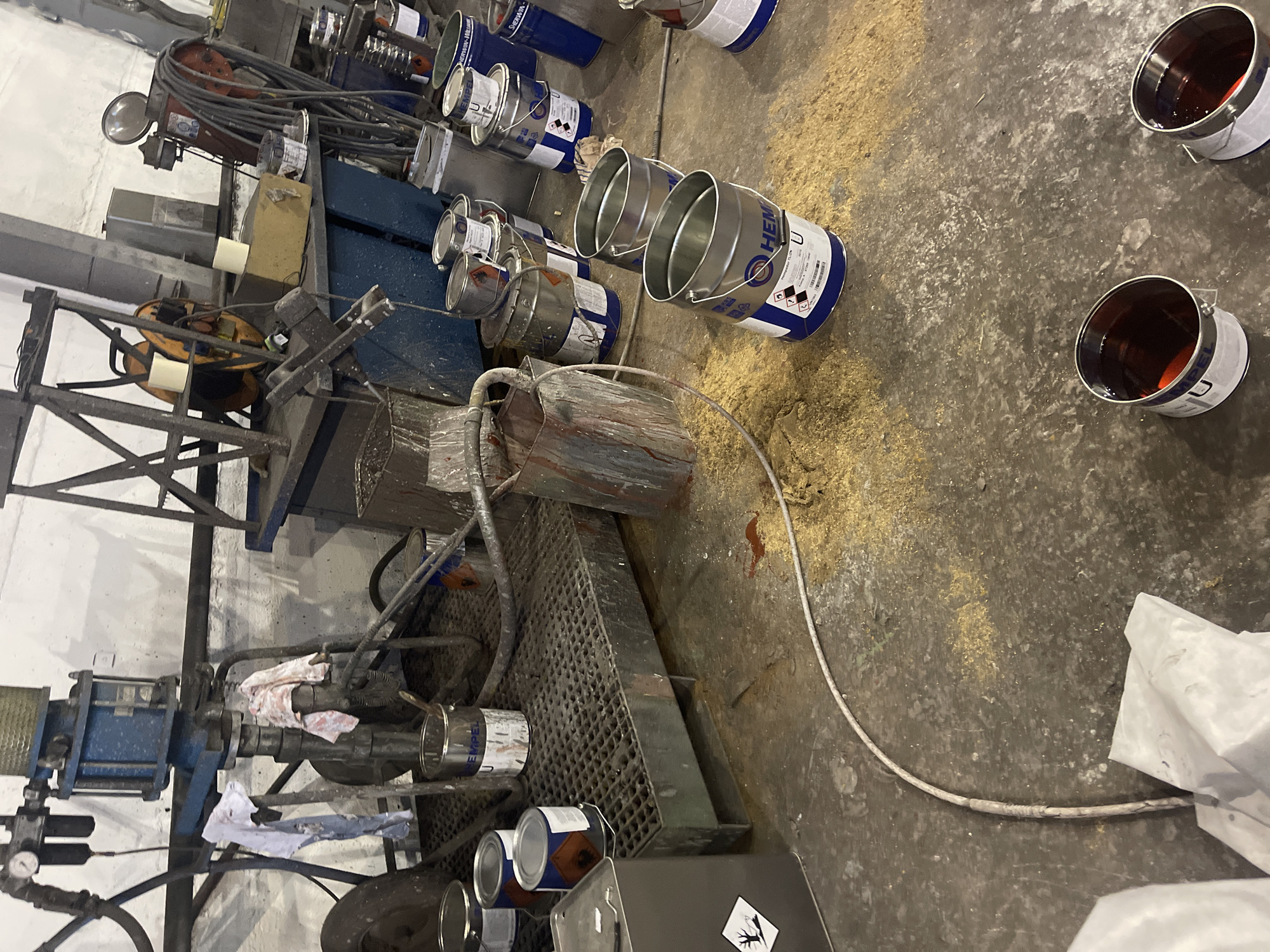 The scene appears to be an industrial workspace or a factory setting in an unknown location. The environment is cluttered with machinery, containers, and scattered materials. There are several metal cans, some of which contain a dark red liquid, potentially paint or a chemical substance. A metal grate or platform is present, along with various pieces of industrial equipment. There is no evidence of people in the image. The overall impression is one of a busy, possibly somewhat disorganized, industrial operation.