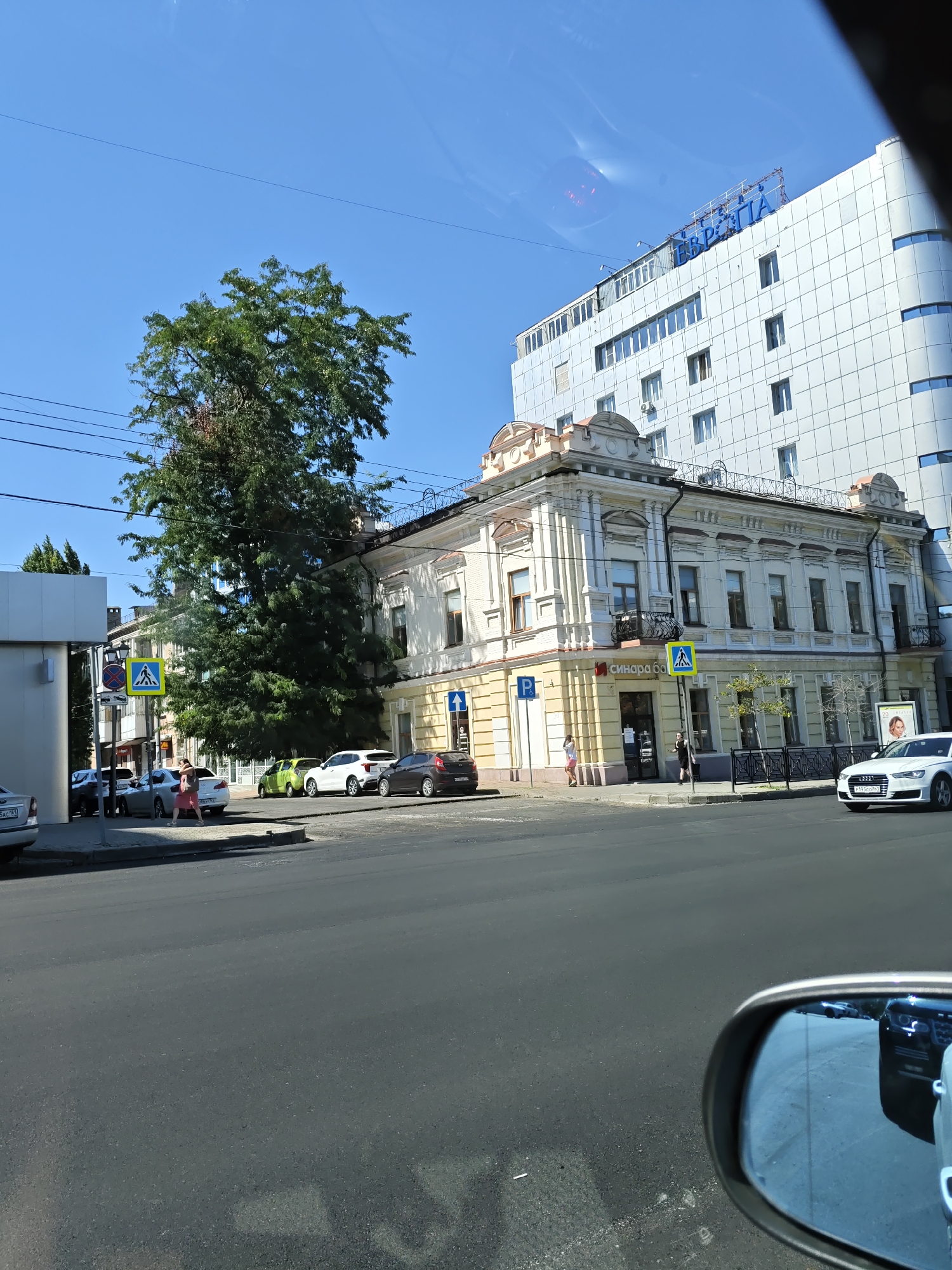 The image captures a sunny day in Rostov-on-Don, Russia, likely in the city center, given the architecture and presence of a large, modern building. In the foreground, a street with cars is visible, and a car's side mirror reflects other vehicles. To the left, a pedestrian crossing is marked on the road, with a yellow pedestrian crossing sign. A tall, green tree dominates the middle ground, casting shadows. Adjacent to the tree is an older, cream-colored building with ornate architectural details and balconies. Behind this building, a large, white, modern building with many windows rises into the clear blue sky. The word "ЕВРОПА" is prominently displayed in blue letters on the top of the modern building. Several cars are parked and driving on the street, and a few pedestrians are seen on the sidewalk. The overall scene depicts a bustling urban environment with a mix of old and new architecture.
