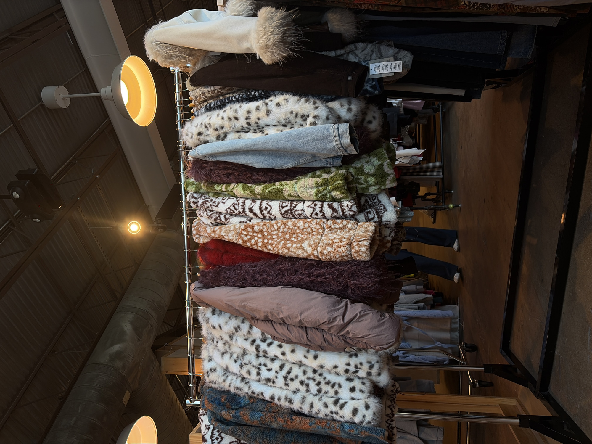 This indoor shot shows a tall, overloaded clothing rack situated in what appears to be a retail space, likely in Austin, United States of America. The rack, standing on wheels, is heavily laden with a chaotic assortment of garments, predominantly jackets or coats with varying textures and patterns. These range from faux fur with spotted prints to solid-colored jackets, possibly made from materials like leather or suede. The rack itself appears to be made of metal with a black frame.
The background reveals a high, industrial-style ceiling with exposed ductwork and lighting fixtures. These features suggest the retail space could be located in a converted warehouse or loft. A concrete floor is seen to the right of the rack.
Hints of other clothing on additional racks can be observed further into the space, including glimpses of jeans and other garments. Feet and lower legs of people shopping or browsing in the store are visible in the background. The scene gives the impression of a busy, possibly vintage or eclectic clothing store with a focus on unique outerwear.