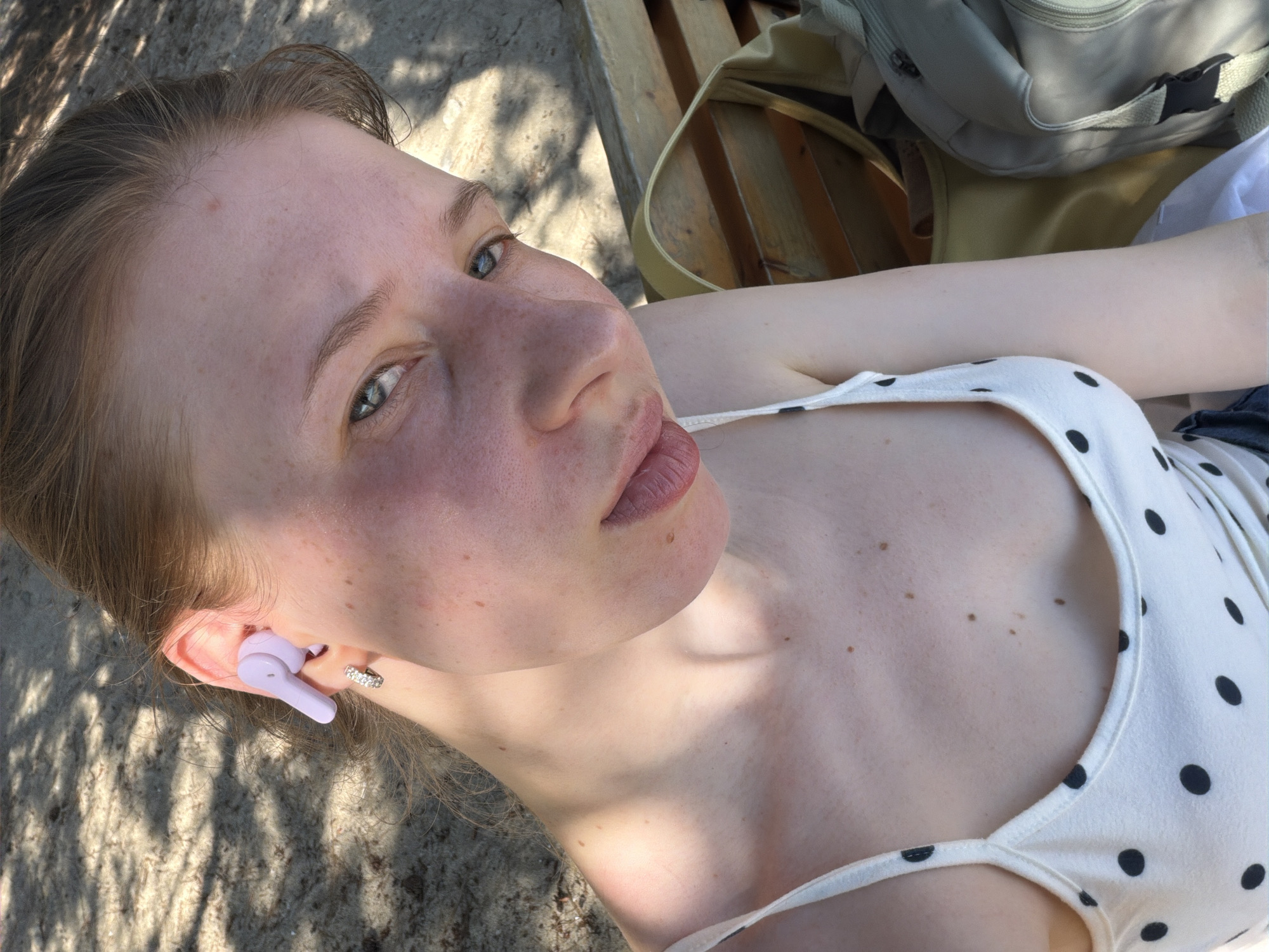 A young Caucasian female, with light skin and freckles, is captured in a close-up selfie outdoors. She has light brown hair pulled back and is wearing a white tank top with black polka dots. A lavender earbud is visible in her left ear, and she is wearing a small hoop earring. Her head is tilted slightly, and she is looking towards the camera. Sunlight casts dappled shadows across her face and shoulders. In the background, the sandy ground and wooden slats of a bench are visible, suggesting an outdoor seating area. A bag is also partially visible to her right. The scene appears to be in a public outdoor space, possibly a park or beach area, in Gangneung-si, South Korea.