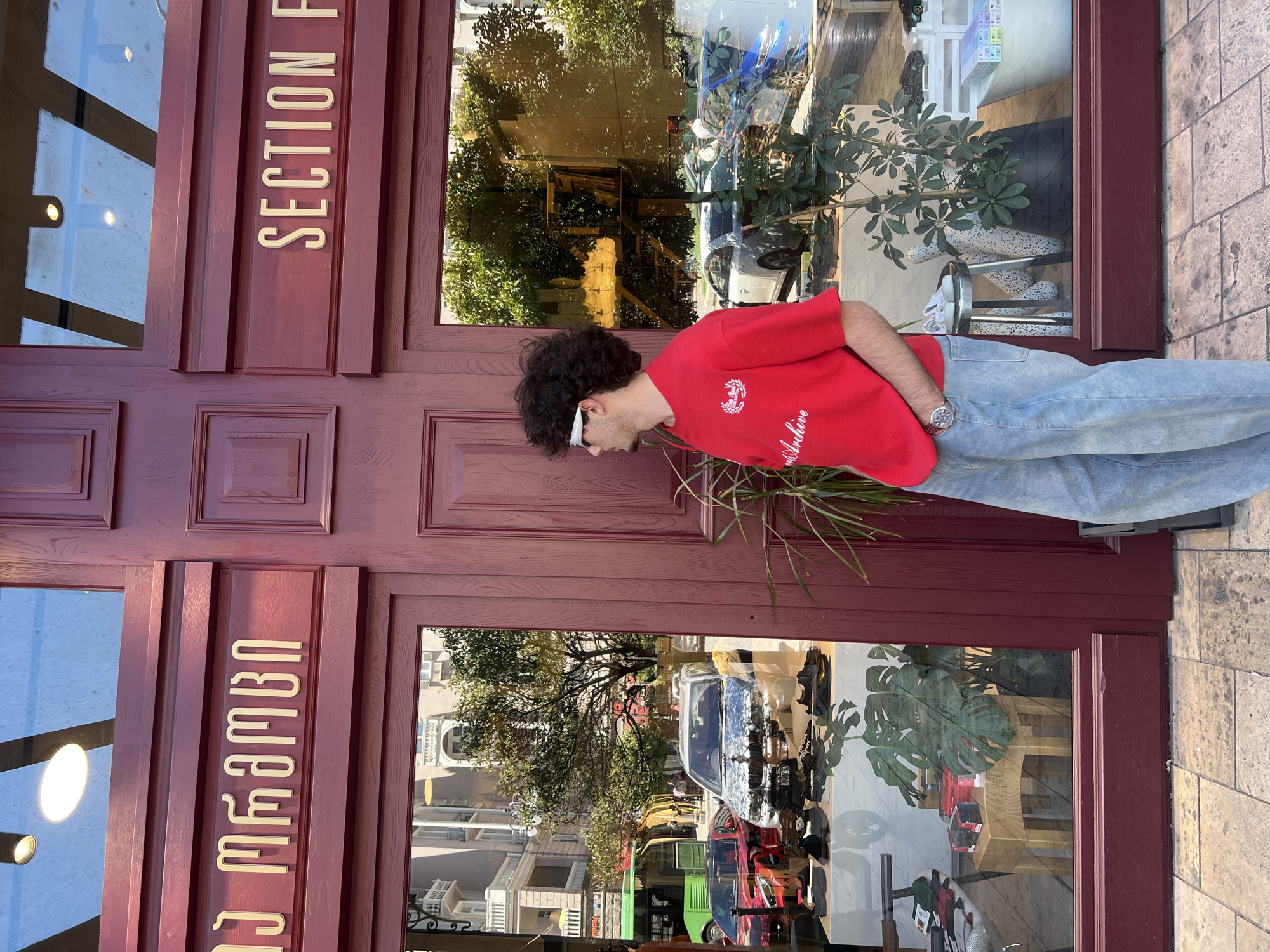A young man with curly dark hair, wearing a bright red t-shirt with white text and sunglasses, stands on a cobblestone sidewalk in front of a storefront in Tbilisi, Georgia. The storefront has a dark red facade with large glass windows. The name of the store is visible in gold lettering on the facade, with Georgian script on the left and "SECTION F" on the right. Through the window, a display of shoes and other merchandise is visible, along with reflections of the street, including cars, a green bus, and buildings. The man is looking to his left, towards the window display, with one hand in his pocket. A potted plant with large leaves stands to the right of the man, partially obscuring the storefront window. The sun is shining, casting shadows and creating bright reflections in the glass.