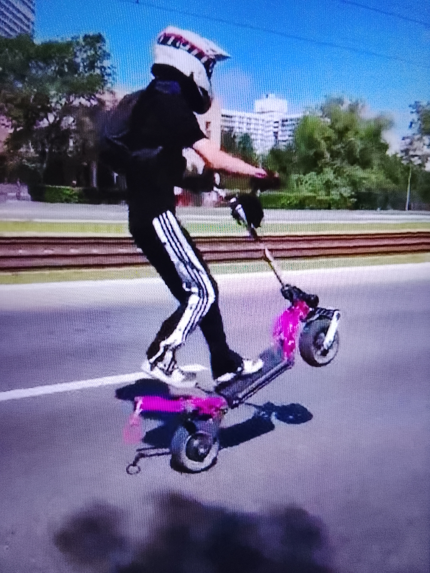 A person wearing a helmet and dark clothing, including pants with white stripes, is performing a stunt on a pink electric scooter. The scooter is airborne, with its front wheel lifted off the ground, suggesting a wheelie or jump. The person is leaning back slightly, balancing on the scooter.
The scene appears to be outdoors during the daytime, under a clear blue sky. In the background, there are trees, buildings, and what appears to be a set of train tracks running horizontally across the image. The setting seems to be urban or suburban.
The image has a slightly pixelated quality, and the colors are somewhat muted, which could indicate a low-resolution photograph or a screen capture. There is no visible text in the image. The activity of the person on the scooter suggests an action sport or recreational activity. The bright pink color of the scooter and the dynamic pose of the rider are the most striking visual elements. The lighting suggests it is daytime.
